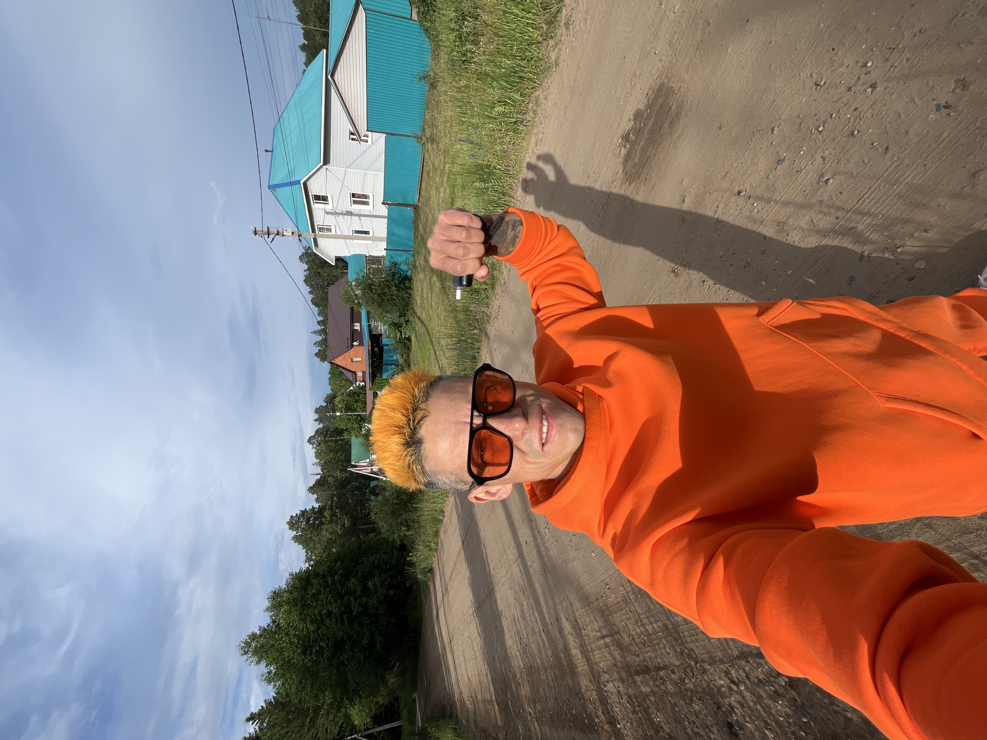 A man with bright orange hair and orange-tinted sunglasses is standing on a dirt road in a rural area. He is wearing an orange hoodie and is holding a black object, possibly a vape pen, in his right hand. In the background, there are houses with brightly colored roofs, trees, and a blue fence. The sky is partly cloudy. The scene appears to be in Chaikovsky, Russia, given the architectural style of the houses and the surrounding vegetation.