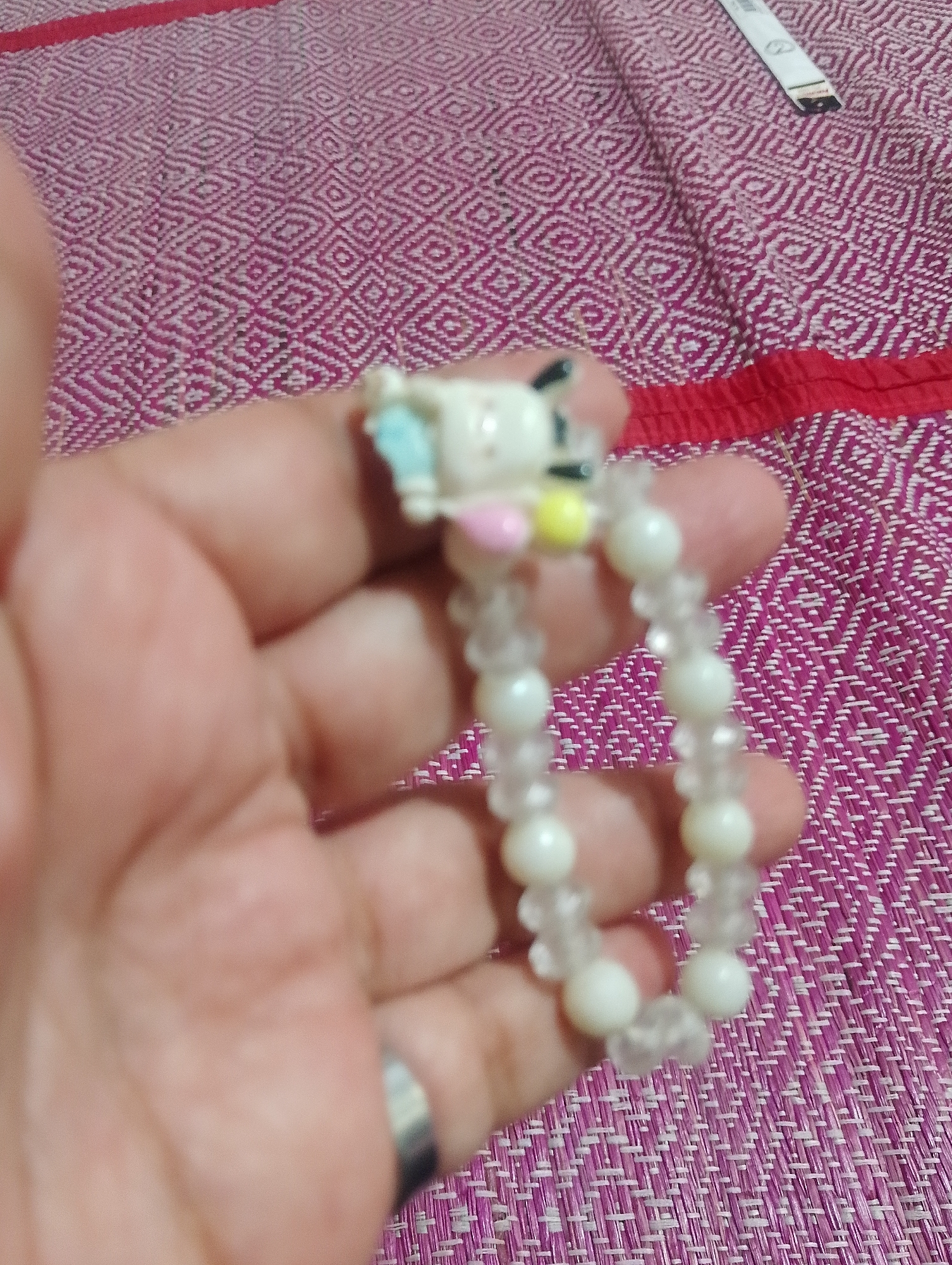 A blurred close-up shot shows a hand holding a bracelet made of white beads and a small cartoon charm. The bracelet is on a textured purple fabric, possibly a mat or textile, with a red stripe along the top edge. There is no discernible urban environment or specific location in Serbelawan, Indonesia.