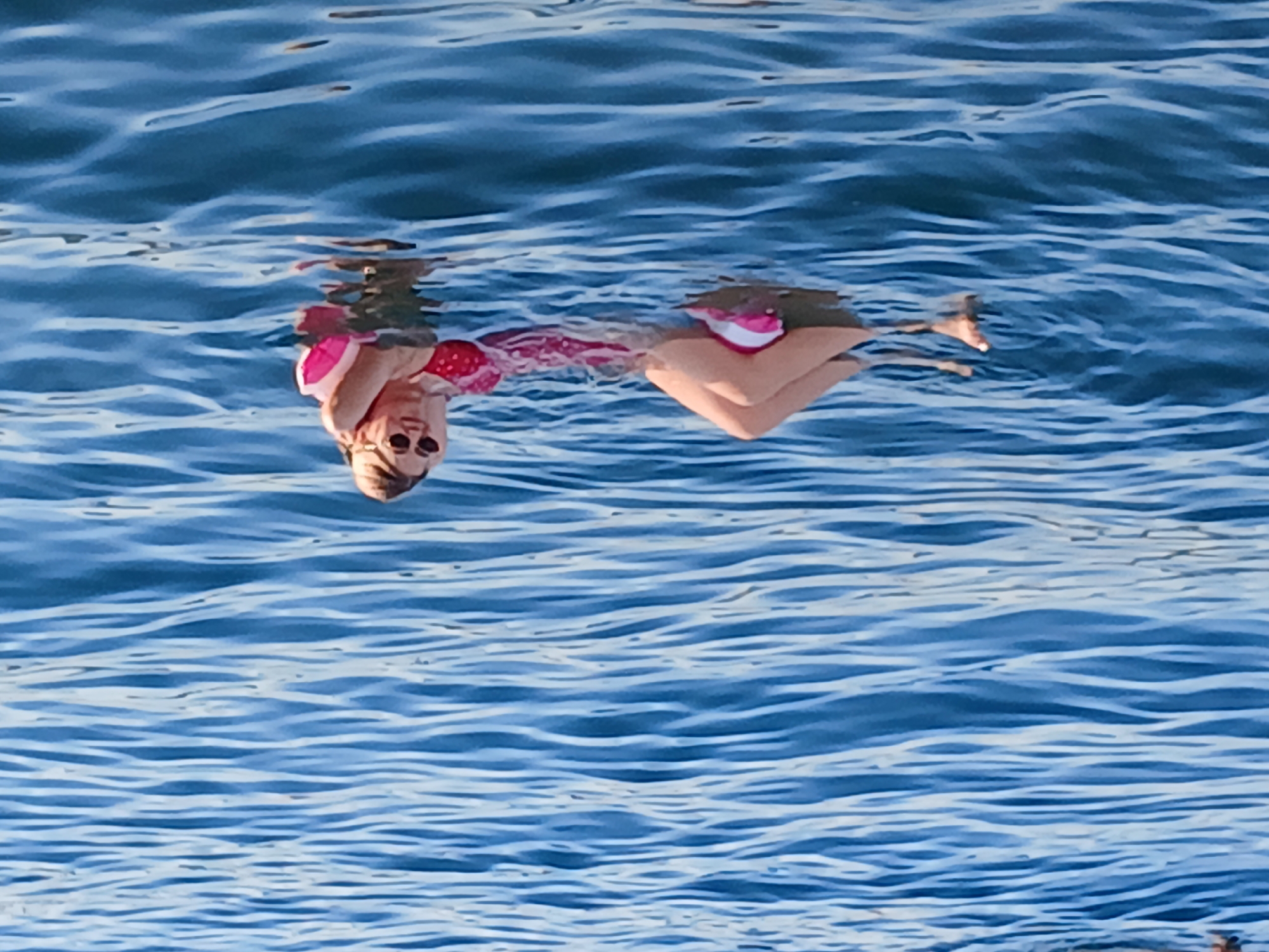 A person is floating on their back in the water. They are wearing a pink and white swimsuit and sunglasses. The water is blue and choppy, with light reflecting off the surface. The scene appears to be a sunny day, and the person seems relaxed as they float. The image is cropped closely to the subject, with the water filling the majority of the frame. The orientation of the image is unusual, with the person appearing upside down.