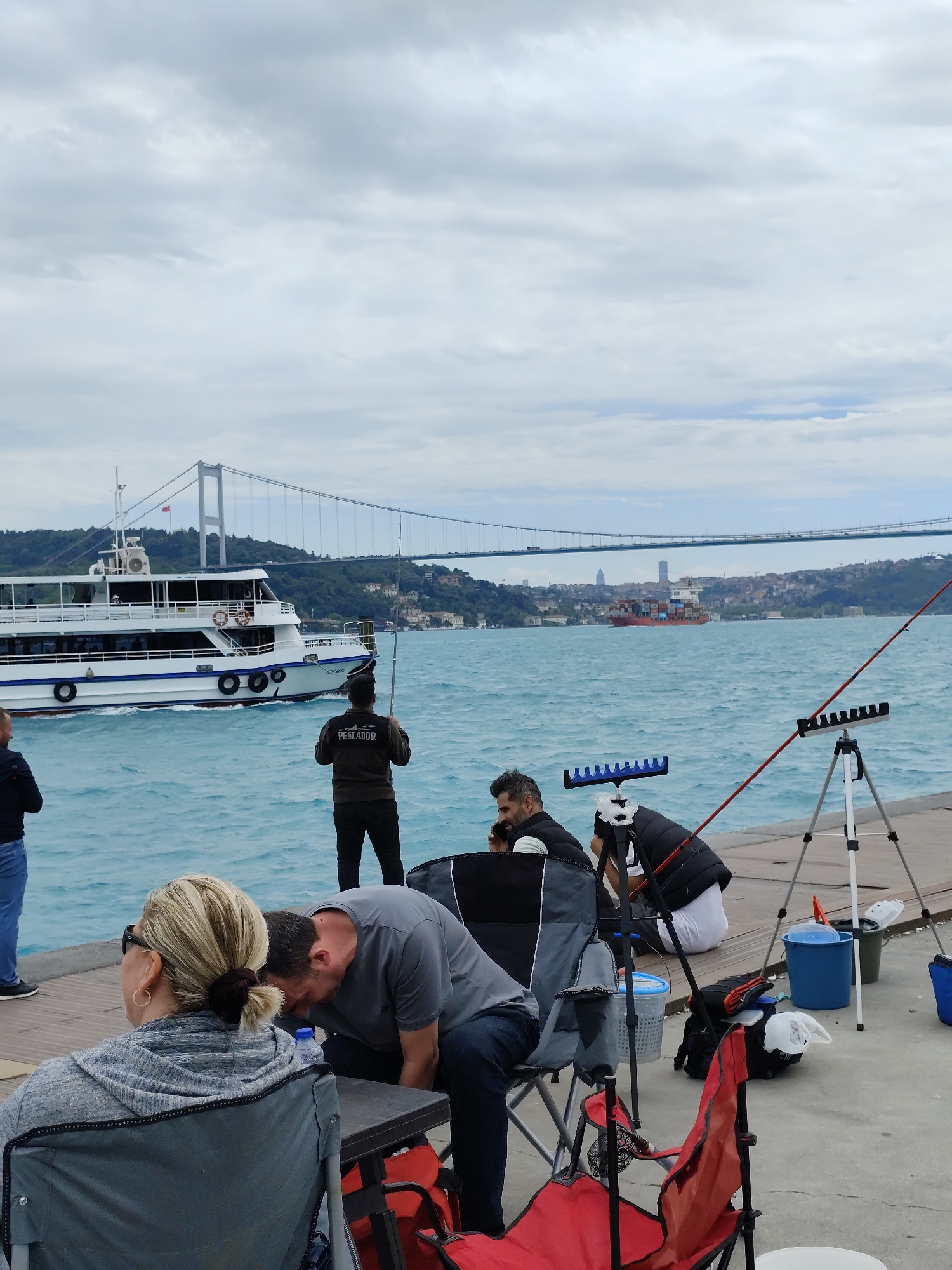 Captured in Sarıyer, Turkey, the image reveals a scene on a wooden pier with people fishing. A ferry with white hull and blue stripes is sailing on a body of turquoise water. The background shows a suspension bridge connecting the land on both sides of the water and the cityscape. The sky is overcast. In the foreground, several individuals are involved in fishing activities, with fishing rods placed in stands. Fishing-related items like buckets, chairs, and tackle boxes are scattered on the pier. Some people are sitting or standing near the fishing gear, while others are looking at the water.