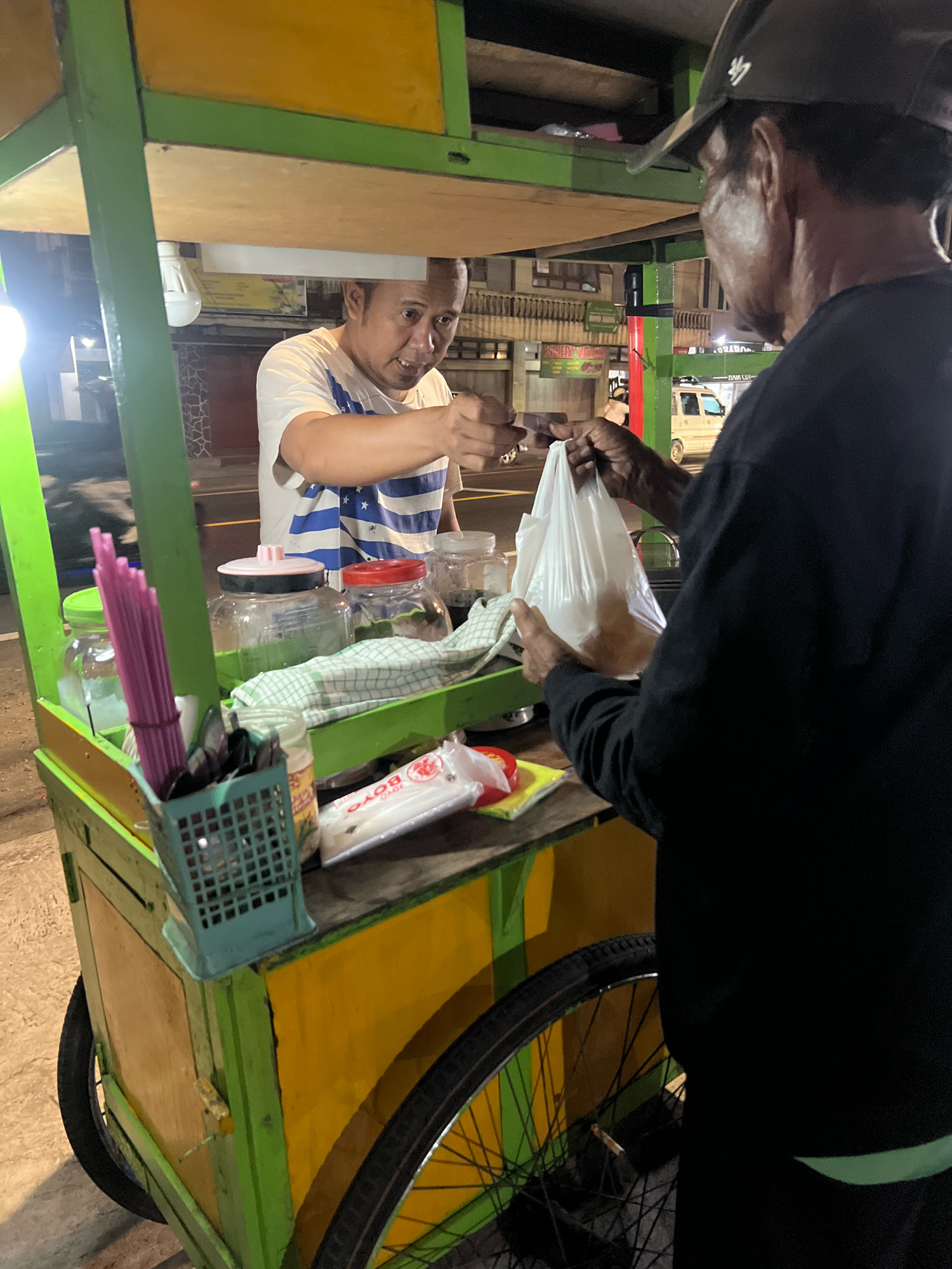 Two men are engaged in a transaction at a street food cart in Payaman, Indonesia. The vendor, wearing a white t-shirt with blue stripes, is handing a white plastic bag containing food to the customer, who is wearing a black long-sleeved shirt and a dark baseball cap. The cart is painted green and yellow, with wooden panels. On the cart's surface, there are several jars, a green basket filled with pink straws and utensils, a checkered cloth, and packaged items. In the background, a street is visible with buildings and a parked vehicle. The lighting suggests it is nighttime.
