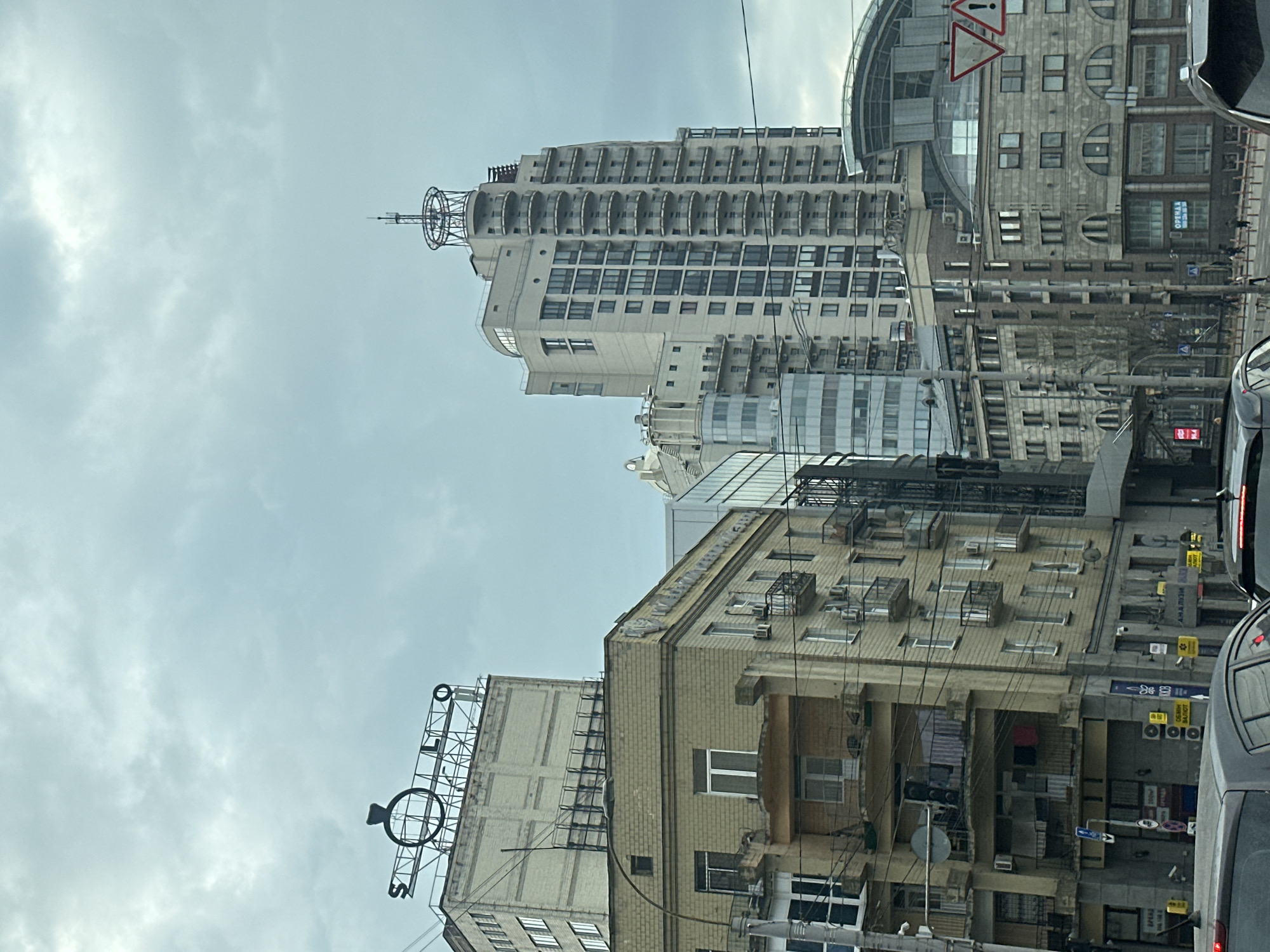 The image captures a street scene in Kyiv, Ukraine, under a cloudy sky. Several buildings dominate the view, showcasing a mix of architectural styles. The tallest building features a distinctive structure on its roof and numerous balconies. Another building bears a vertical sign with the word "SOLO," along with a large ring symbol. Overhead utility lines are visible, crisscrossing the scene. A few vehicles are present on the street, suggesting traffic activity. People are seen walking on the sidewalks next to the buildings.
