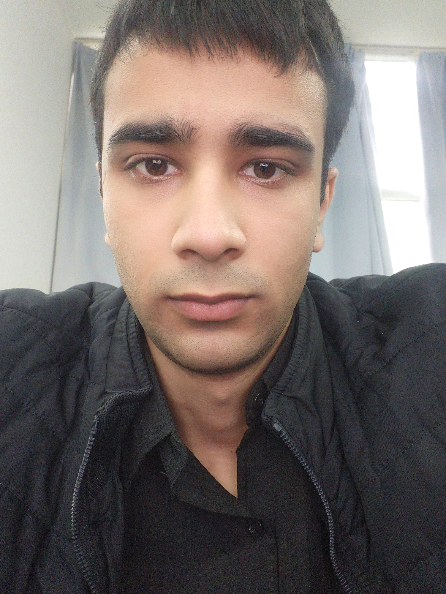 A close-up selfie of a man with dark hair and brown eyes. He is wearing a black quilted jacket over a black shirt. The background includes a window with light coming through and gray curtains. There is no evidence in the image to determine the location is in the city of Chongqing High-tech Zone, China.