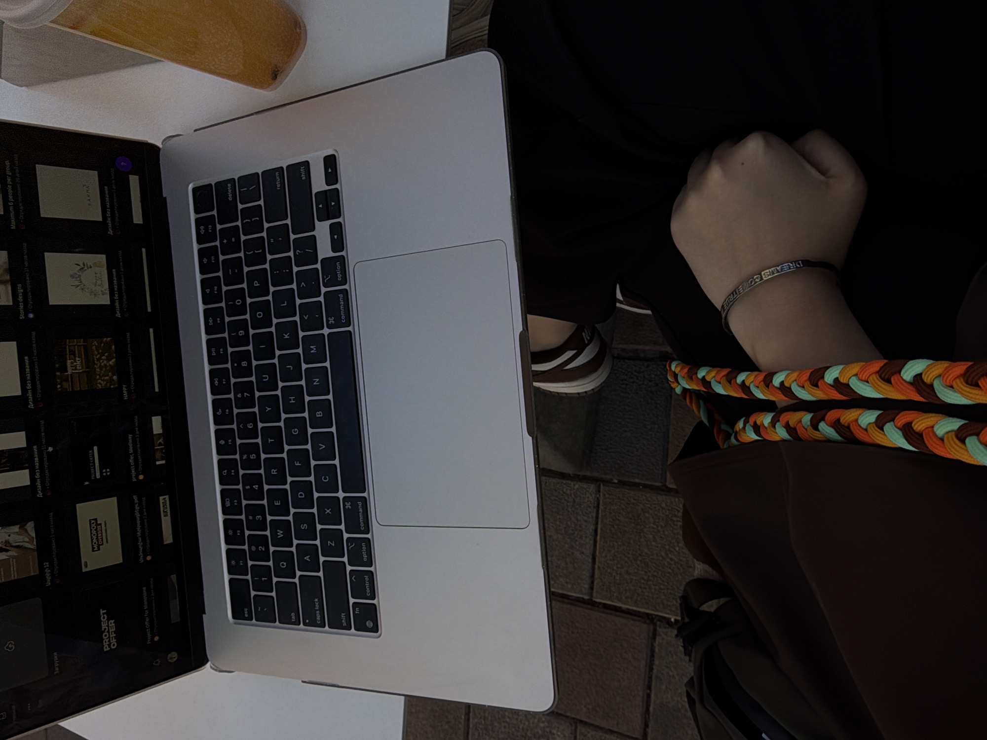 A person sits outside with a laptop, presumably working. The laptop displays a design project with options in Armenian. The person has a bracelet on their wrist that says "Dreams Come True" and a colorful braided strap of a bag or purse sits on their lap. They are wearing dark pants and a brown jacket. The location is likely Yerevan, Armenia, based on the Armenian text on the laptop screen. The setting is urban, possibly a cafe terrace, indicated by the brick pavement visible near the person's shoe.