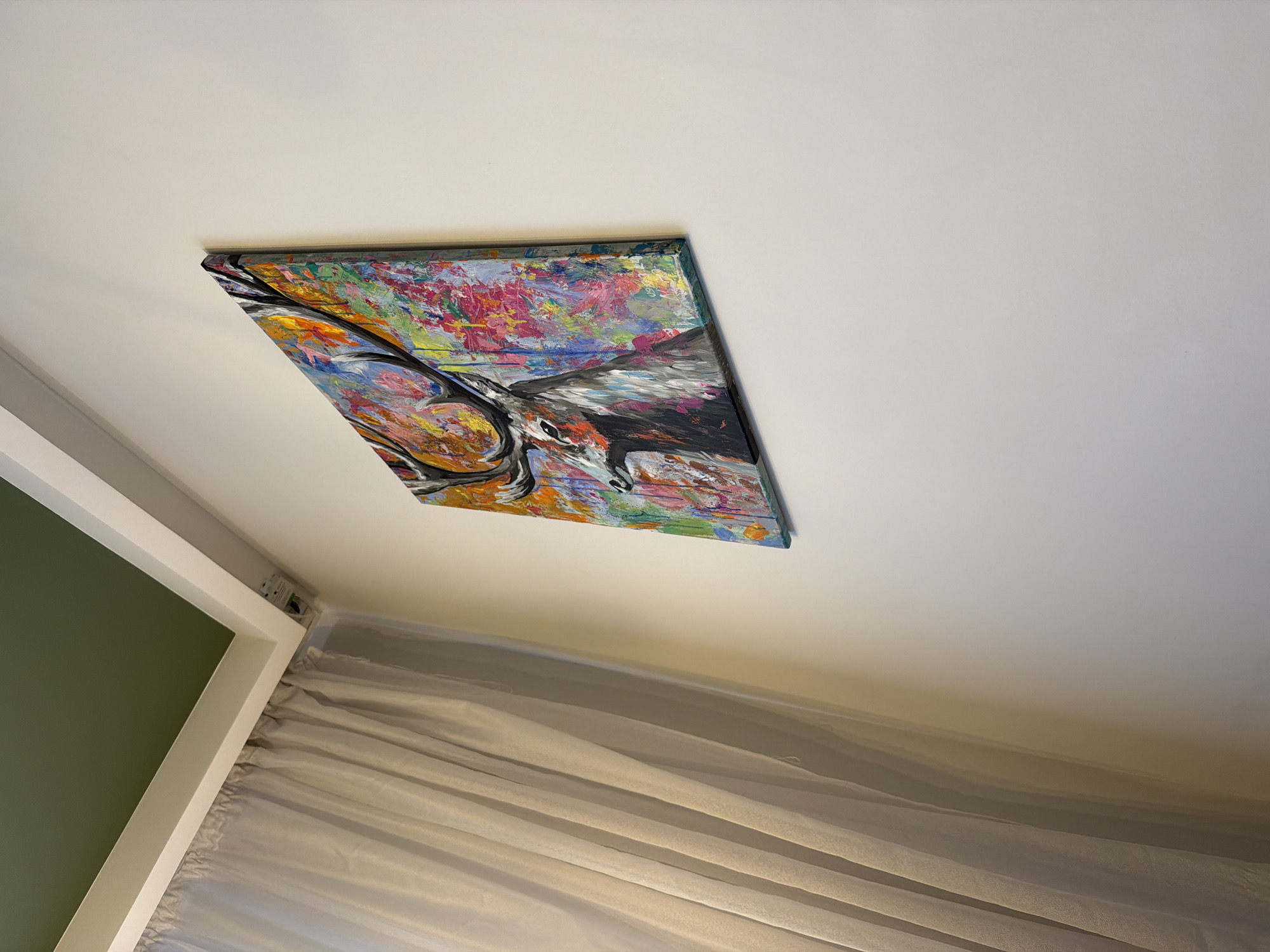 The image depicts an interior scene, likely a room, featuring a canvas painting of a stag with vibrant, abstract colors. The painting is mounted on a plain white wall. To the left of the painting, sheer, light-colored curtains are visible, which appear to be drawn back. Above the curtains and the wall, a section of the ceiling is visible, painted in a dark green hue, with white trim. The perspective is from a low angle, looking upwards towards the ceiling and the right side of the room. There are no people or animals present in the scene, and no specific details suggest a location in Brest, Belarus. The scene appears to be a still, decorative interior.