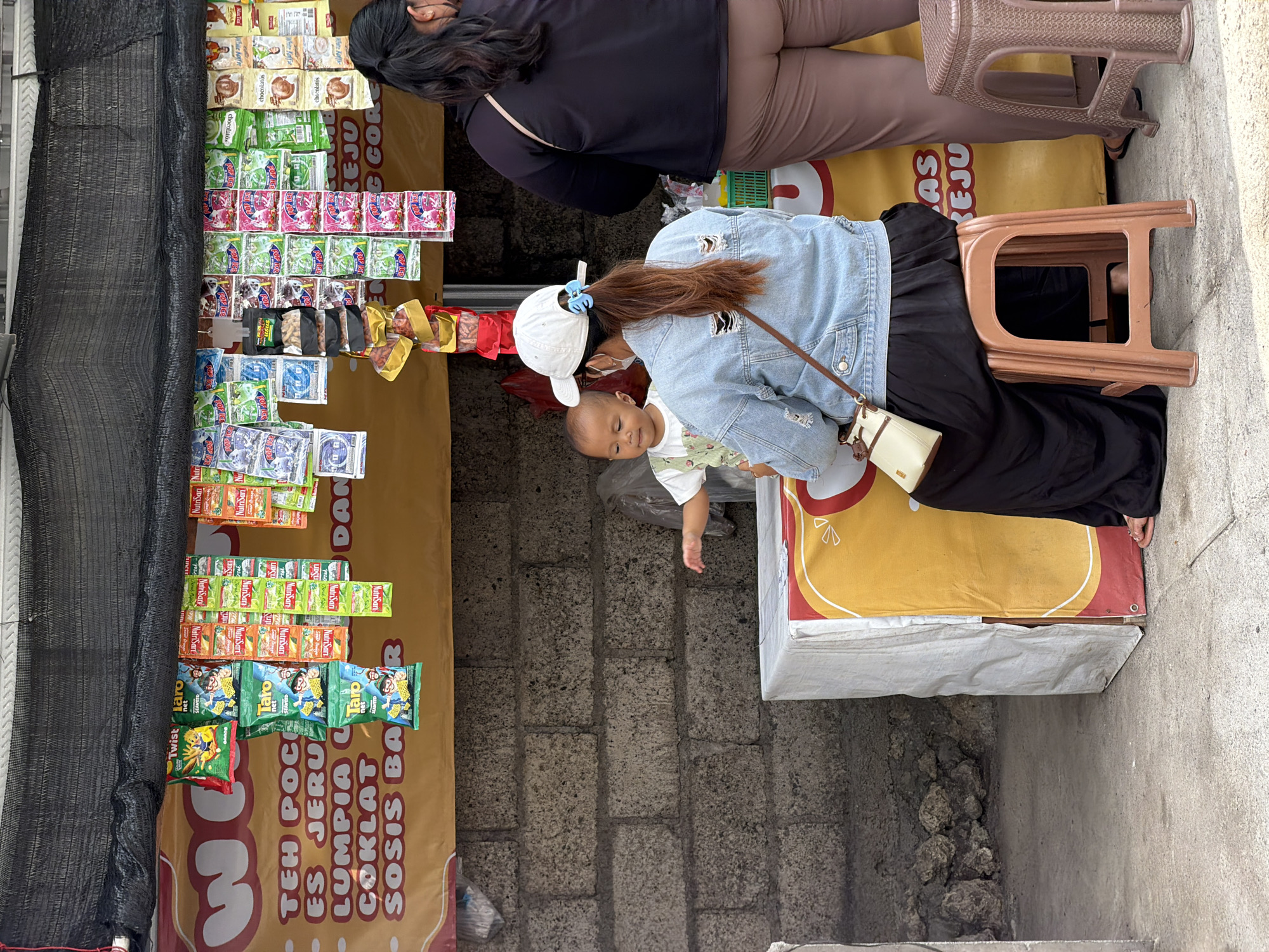 A medium shot captures a street vendor's stall, possibly in an outdoor market or roadside setting. The stall is covered by a black mesh tarp and displays a variety of packaged snacks and drinks hanging from the front. A yellow banner with Indonesian text is draped across the front of the stall, listing various food items like "TEH POC", "ES JER", "LUMPIA", "COKLAT", and "SOSIS BA".
In the foreground, a woman wearing a blue denim jacket, a white baseball cap, and a black skirt is seated on a brown plastic stool. She has a white crossbody bag with a tan strap. Next to her, a young child in a light-colored shirt is standing and reaching out towards the stall.
To the right, another woman in a black top and brown pants stands, facing away from the camera, partially obscuring the stall's display. A brown plastic stool is visible to her right. The background consists of a rough, gray brick wall and a concrete ground surface. The overall scene suggests a casual commercial interaction.