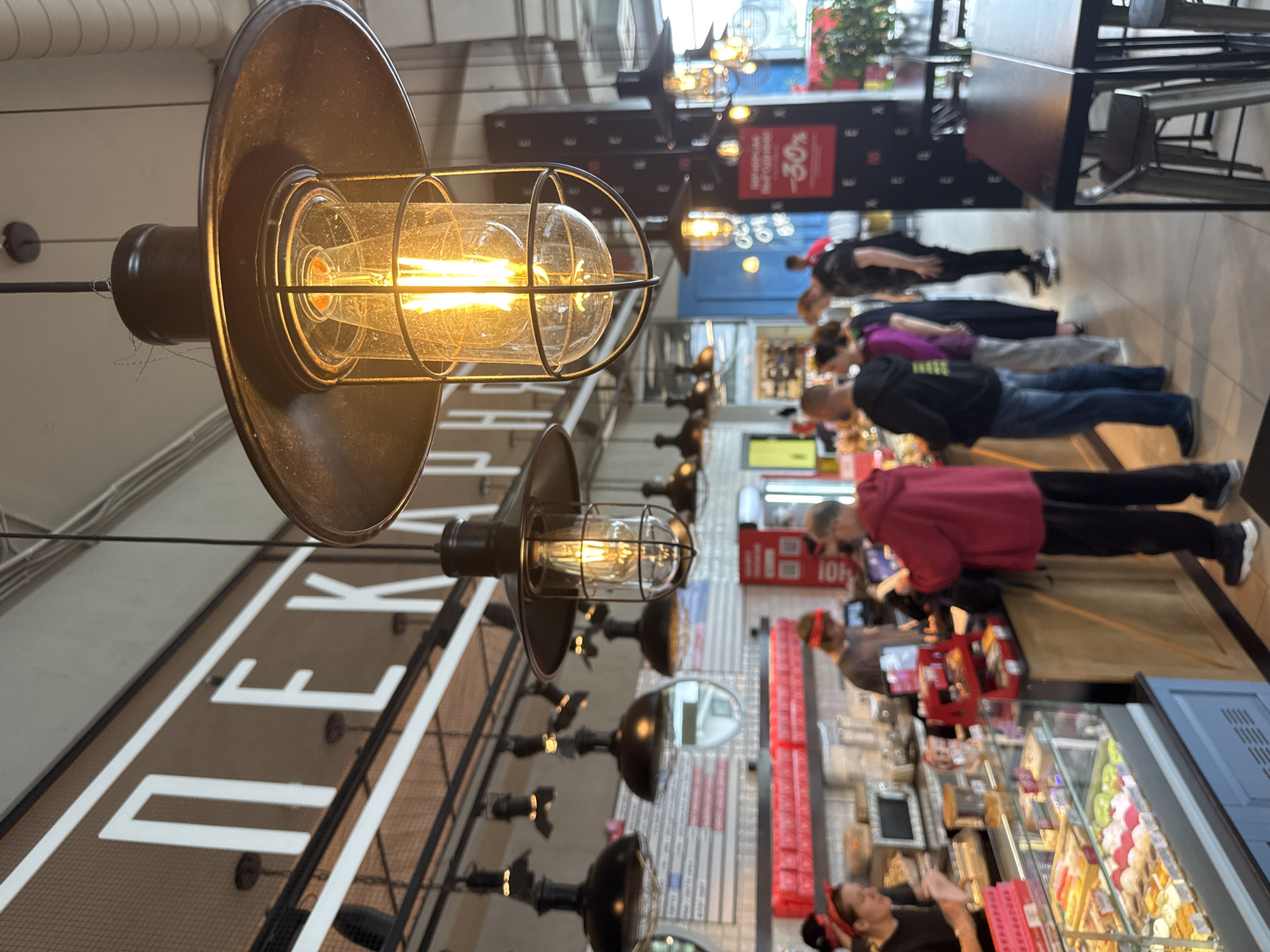 This image depicts the interior of a food market or a similar establishment, likely a cafe or a food hall, with a focus on the overhead industrial-style lighting. Several people are visible in the background, standing in a line, suggesting they are customers waiting to be served. One person in the foreground appears to be interacting with a food display case. The overall lighting is warm and inviting, likely due to the incandescent bulbs in the prominent light fixtures.
The scene is populated with various objects, including display cases filled with food items, tables, and signage. The architectural style suggests a modern, possibly trendy, setting. The people in the image are dressed casually, indicating a daytime activity. Reflections in the polished floor and surfaces add depth to the scene.
The visible text includes "30%" on a red sign in the background, possibly indicating a sale or discount. Other text is partially visible on signage behind the customers, but it is not legible enough to decipher. The overall impression is one of a busy commercial space, likely during operating hours. The camera angle is from a low perspective, looking up towards the lights and then downwards towards the people and displays. The image has a somewhat tilted orientation, adding a dynamic feel to the composition.