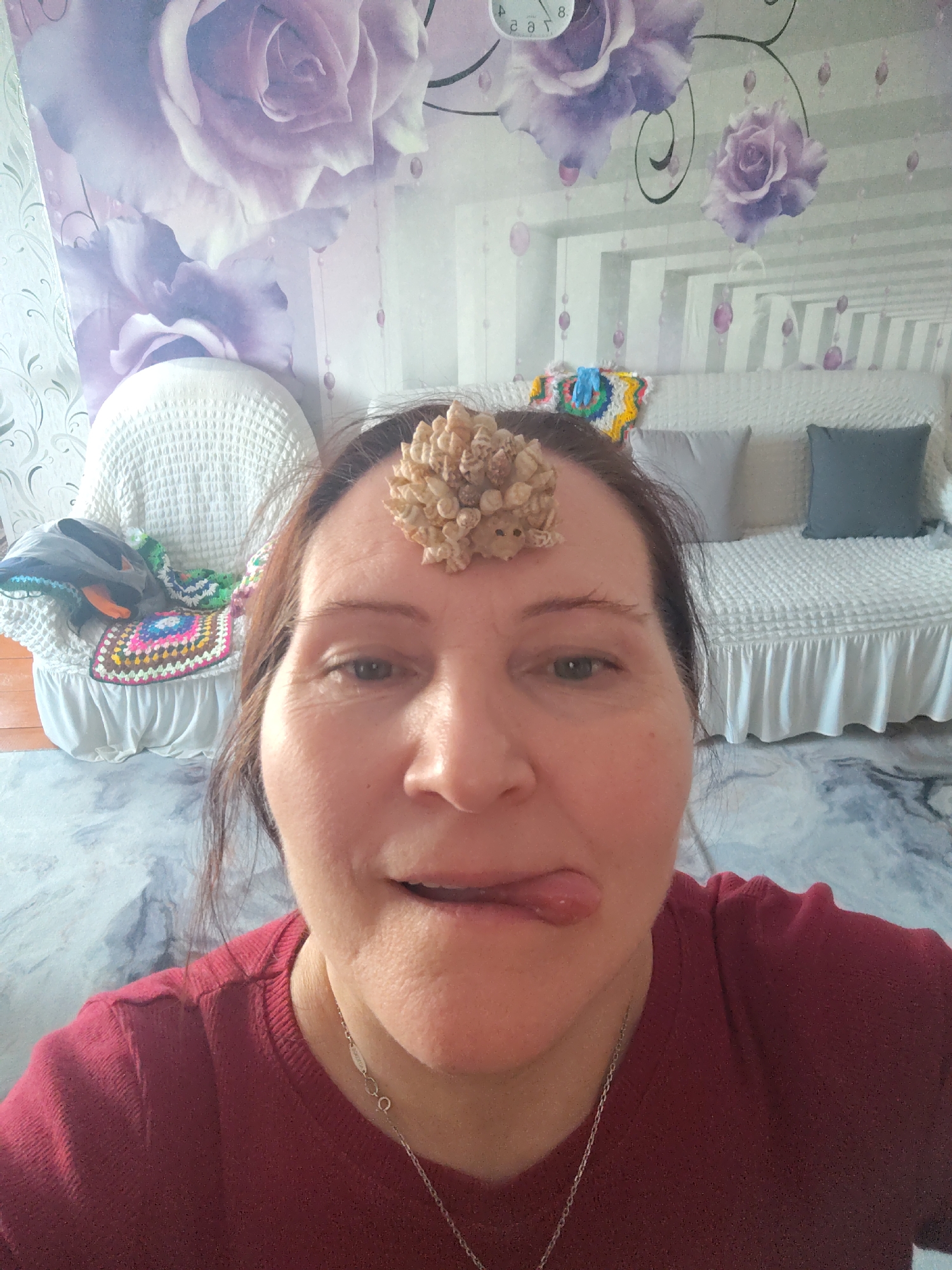 A medium-shot portrait captures a middle-aged woman indoors in Сусады-Эбалак, Russia. She looks directly at the camera with a playful expression, her tongue slightly extended to the side of her mouth. A cluster of small, light-colored, irregular objects, possibly shells or dried botanical elements, is balanced on her forehead. She has brown hair pulled back and is wearing a red long-sleeved shirt and a silver chain necklace.
The background reveals a domestic setting, likely a bedroom or living room. The wall is covered in wallpaper featuring large, stylized purple roses, green leaves, and decorative vine-like elements with small, translucent purple bead-like drops. In the upper left corner, part of a clock showing "8:" is visible, appearing reflected.
To the woman's left, a white, woven armchair is visible, adorned with a colorful crocheted blanket or throw and other fabrics. To her right, a white bed or sofa with a ruffled base is covered in a textured white bedspread. Two pillows, one light grey and one dark grey, rest on the bed/sofa. The floor is covered by a patterned rug or carpet in shades of blue, grey, and white, resembling abstract marble. The overall impression is casual and personal, with a sense of whimsy due to the woman's pose and the object on her forehead.