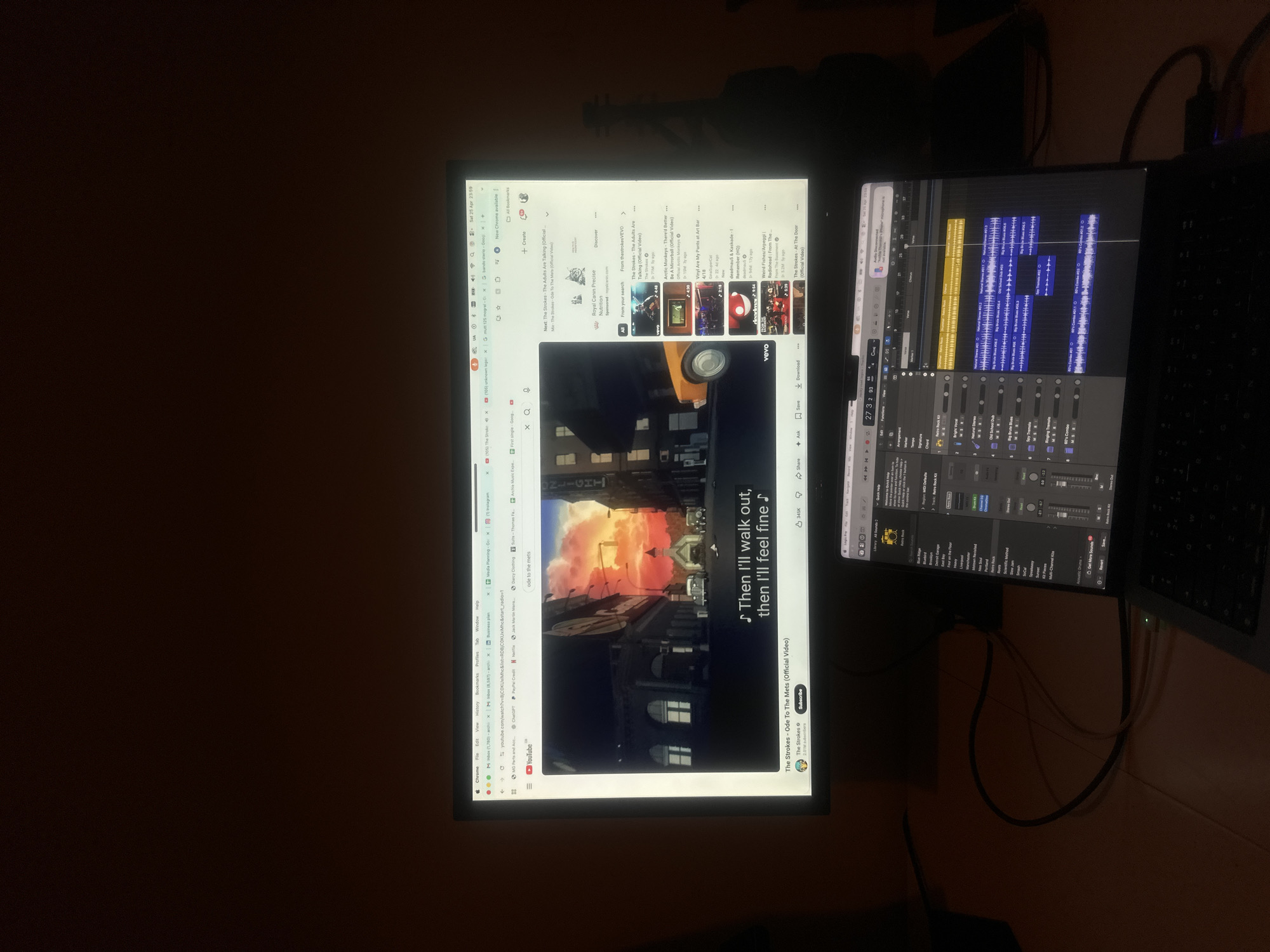 The image shows two screens: a larger monitor on the left and a laptop screen on the right.
The larger monitor displays a YouTube video of The Strokes' "Ode To The Mets." A web browser is open with multiple tabs, including search queries like "mutt 125 mogral," "First single Google" and "ChatGPT." Bookmarks related to music production and other topics are visible.
The laptop screen displays a digital audio workstation (DAW) interface, likely Logic Pro. The DAW shows waveforms of audio tracks arranged in a timeline, suggesting the user is working on a music production project.
A black object, possibly a guitar, sits in the background above the monitor. A dark room encloses this space.
I cannot identify the specific location in London, United Kingdom.