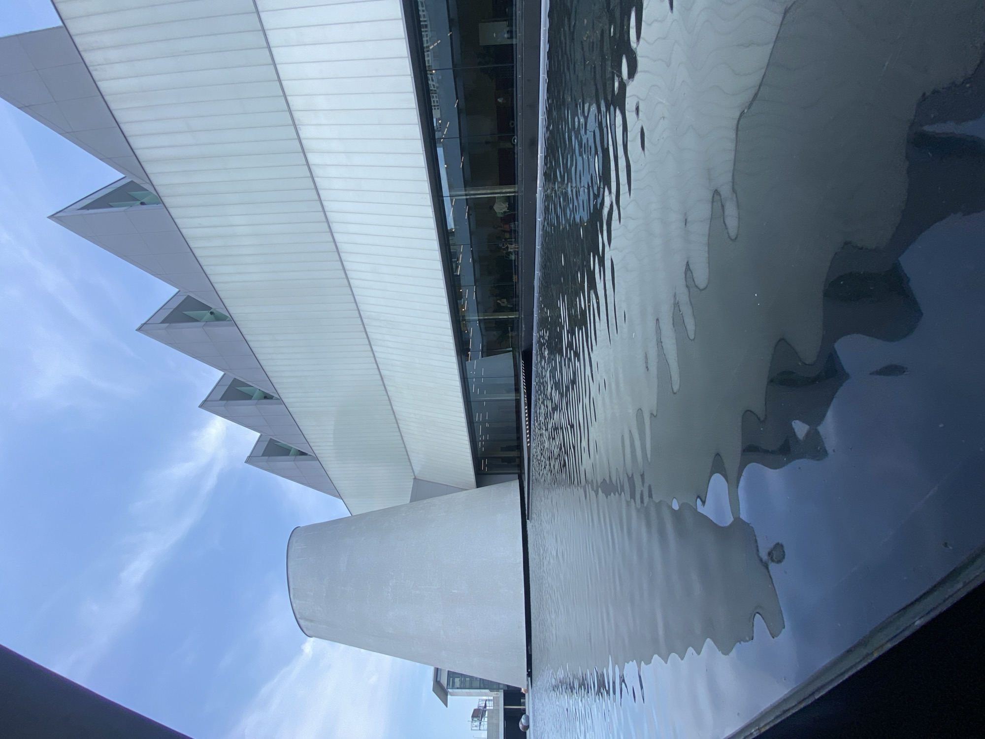 The image shows a modern building reflected in a body of water, likely a pool or reflecting pond. The building is characterized by a predominantly white facade with angular, triangular extensions that create a distinctive sawtooth pattern. Glass windows are visible, reflecting the interior. Part of the building is cylindrical, possibly a supporting structure or architectural feature. The sky is pale blue with scattered clouds. The water's surface is rippled, distorting the reflected image of the building. The photograph's perspective is angled, giving a dynamic, upward-looking view of the scene. The location is the Bangkok Art and Culture Centre (BACC), a contemporary arts centre in Bangkok, Thailand.