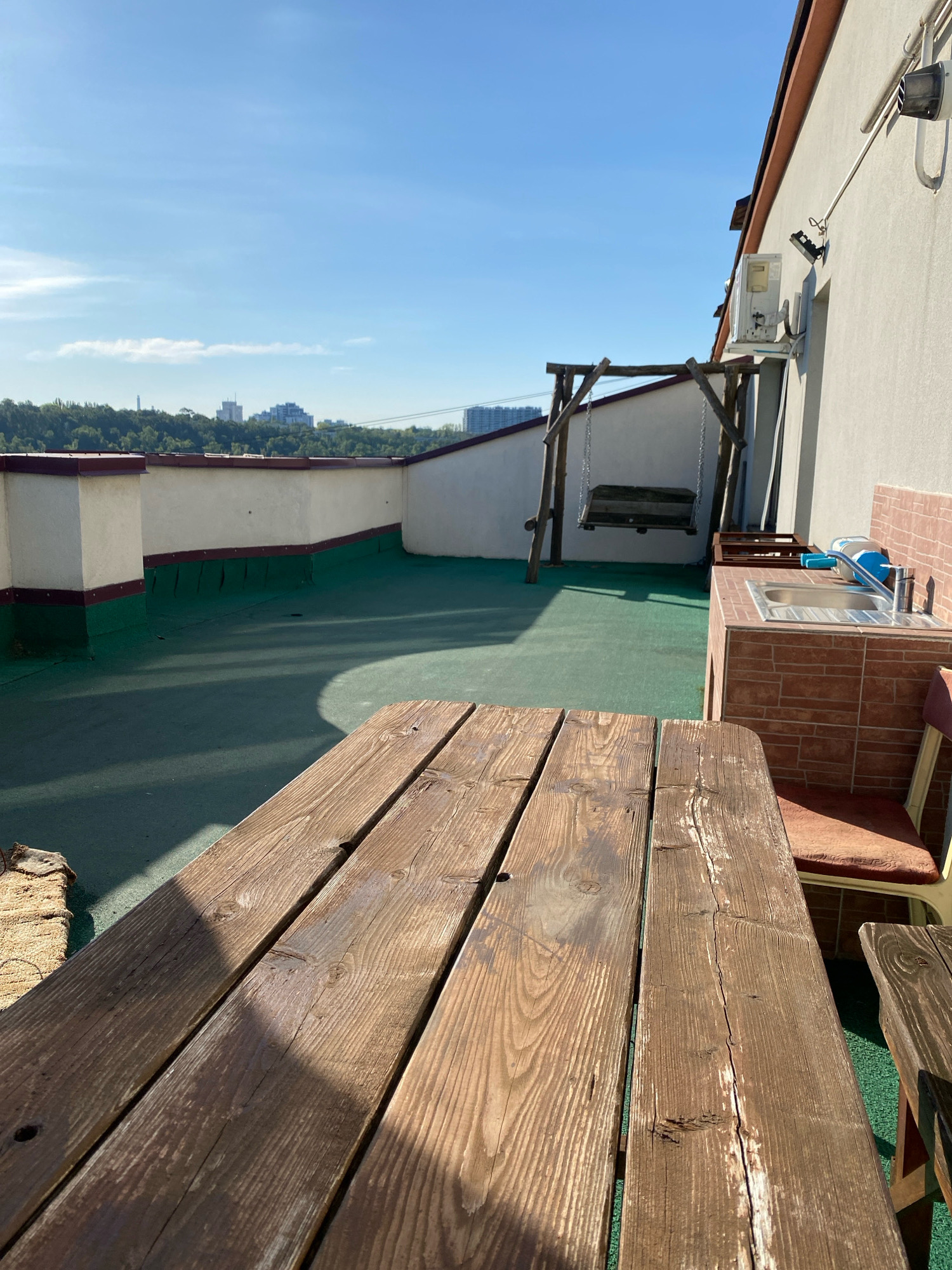 The image depicts an outdoor rooftop terrace with a clear blue sky and distant city buildings visible in the background. The terrace is covered with a green surface, and there is a wooden picnic table in the foreground. To the left, a white wall with a brown trim extends along the edge of the terrace, with a dark green section beneath it. A wooden swing set with a bench is situated further back. On the right side of the image, there is a sink area with a metal basin and a chrome faucet, built with brick-like tiles. An air conditioning unit is mounted on the exterior wall. The overall scene suggests a recreational outdoor space. The specific location in Chișinău, Moldova, cannot be determined from the visual information provided in the image.