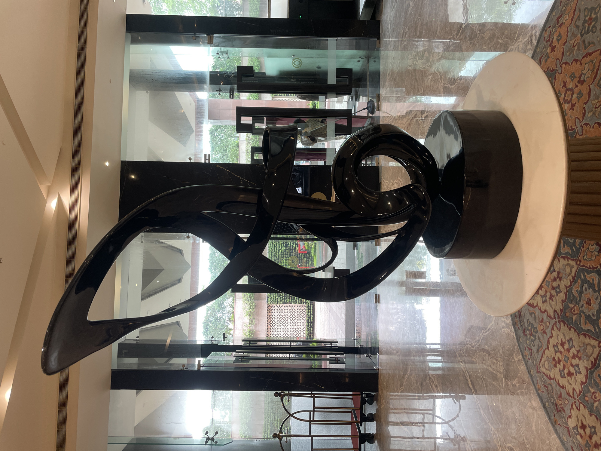 The image shows a large, abstract black sculpture displayed in the lobby of a hotel in Agra, India. The sculpture, made of a glossy material, is positioned on a circular white pedestal which rests on a round rug. The lobby features polished marble floors that reflect the natural light streaming in from the glass doors and windows. Behind the sculpture, large glass doors lead to an outdoor area with greenery and patterned walls. To the left, a luggage cart is visible. People are seen through the glass doors, suggesting the location is a public or semi-public space. The overall impression is one of modern elegance within a grand interior.