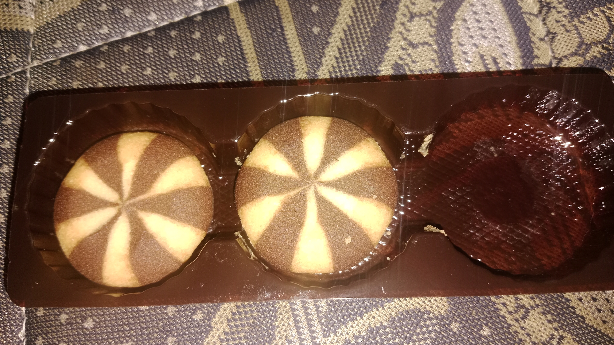 A package of three chocolate and vanilla swirl cookies and one dark chocolate tart is shown in a brown plastic tray. The tray is placed on a textured grey and beige fabric. The cookies are round with alternating stripes of chocolate and vanilla. The tart appears to be filled with a dark, viscous liquid. The image is a close-up, overhead shot.
