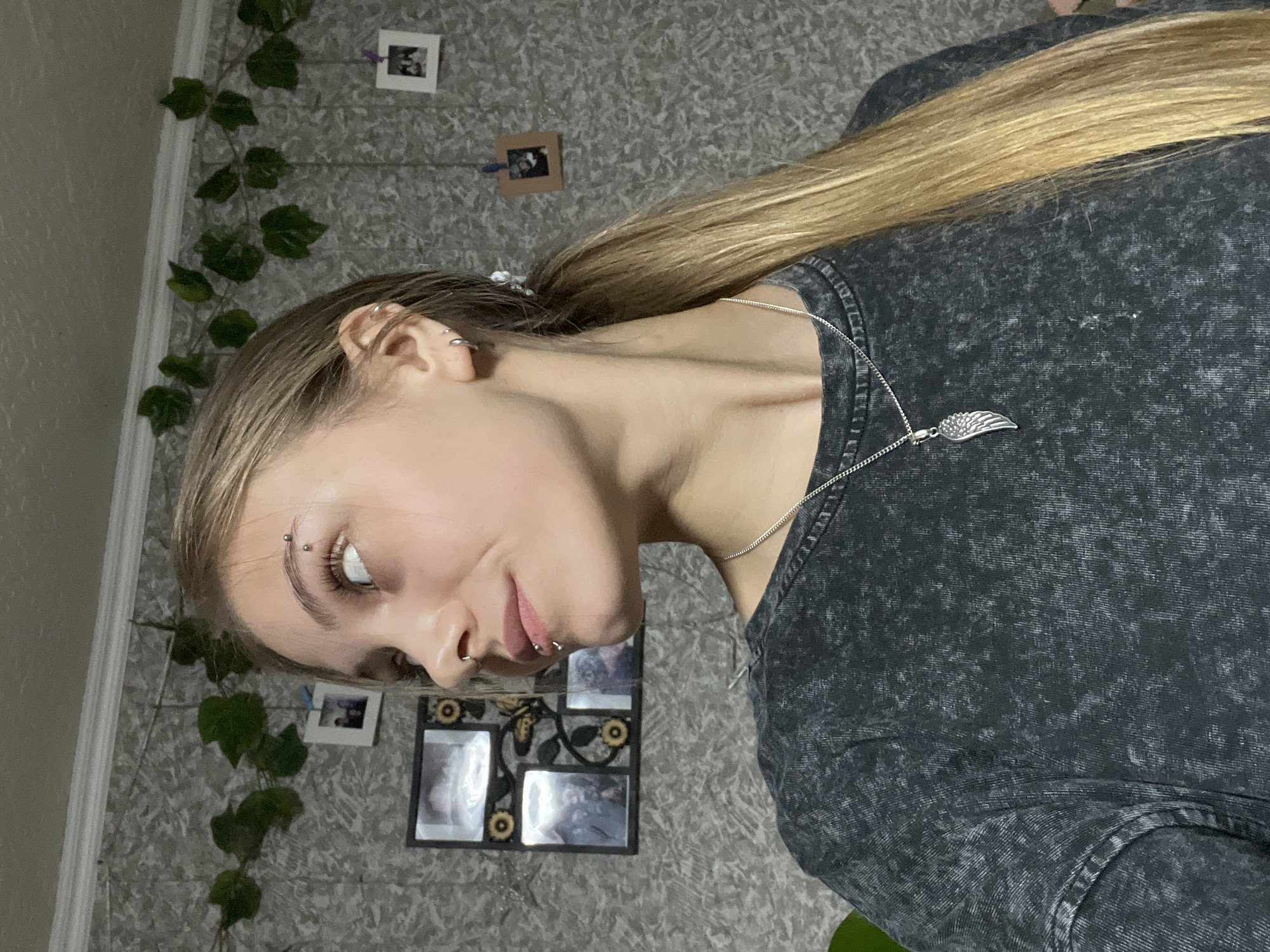 The image depicts a young female, appearing to be in her late teens or early twenties, presented in a chest-up portrait format, set indoors in a room in the city of Фоки, Russia. She is turned slightly to her left, with her head tilted back and her gaze directed upwards and to the right. Her long, straight, blonde hair is pulled back into a low ponytail, resting over her right shoulder.
Her left facial profile is visible, showcasing several piercings: a double-barbell piercing on her left eyebrow, a silver septum ring through her nose, and two small silver studs indicative of vertical labret piercings on her lower lip. Her left ear is adorned with multiple silver hoop earrings. She wears a dark grey, long-sleeved garment with an acid-wash or stonewash texture. A silver chain necklace with a detailed wing-shaped pendant is visible around her neck.
The background wall features a textured grey wallpaper with a speckled pattern. A white crown molding runs along the top of the wall, from which artificial green ivy-like leaves are draped. Several small, square photographs or polaroids are pinned to the wall, some secured with small clips (one purple clip is visible). To the left of the subject's head, a multi-section black picture frame, embellished with circular golden accents and a butterfly motif, hangs on the wall.