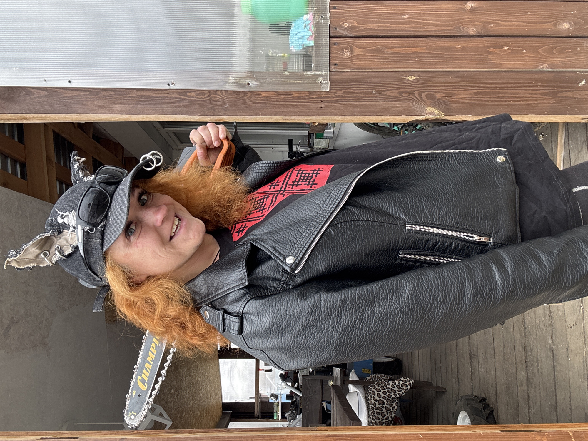 A medium shot depicts a woman with fair skin, red hair, wearing a black leather jacket, a black t-shirt with a red graphic, and a gray cap with goggles, seemingly indoors. She holds a gray chainsaw with the brand "Champion" visible on the blade. The background includes a wooden structure with corrugated plastic, and various items such as a tractor tire and a leopard-print cloth partially visible. The location is unspecified but believed to be Chaikovsky, Russia.