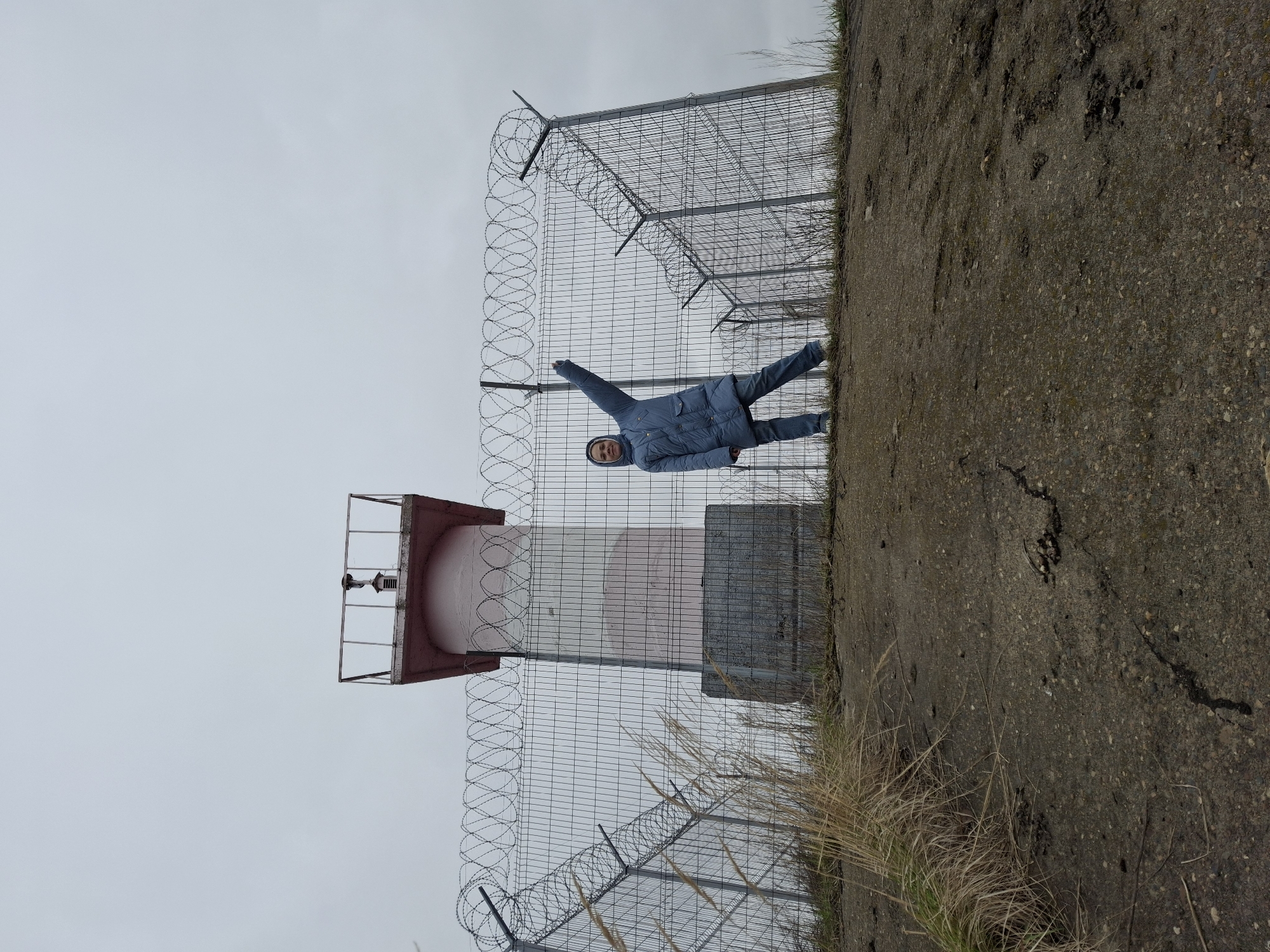 The image depicts a person in a blue hooded jacket standing next to a red-and-white lighthouse-like structure surrounded by a wire fence topped with barbed wire. The location is Chaikovsky, Russia. The person is facing the camera with one arm raised. The background consists of a gray sky and a concrete surface covered with grass. The scene appears to be taken from a low angle, with the lighthouse structure and fence towering over the ground level.