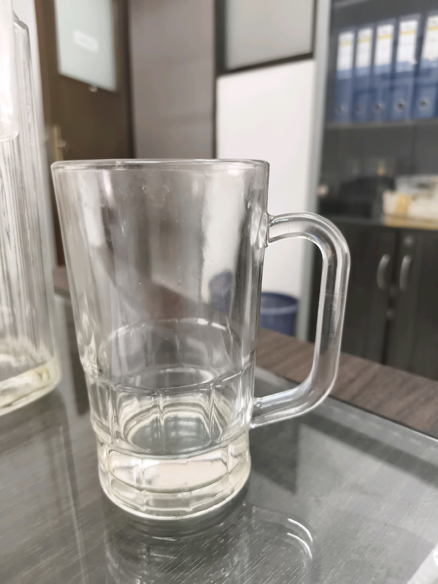 An empty, clear glass mug with a handle sits on a reflective, dark surface. The mug has a fluted base and a straight rim. In the background, out of focus, a filing cabinet filled with blue binders is visible. To the left, a portion of a tall, ridged glass container is also present. The scene appears to be an indoor office or storage area.