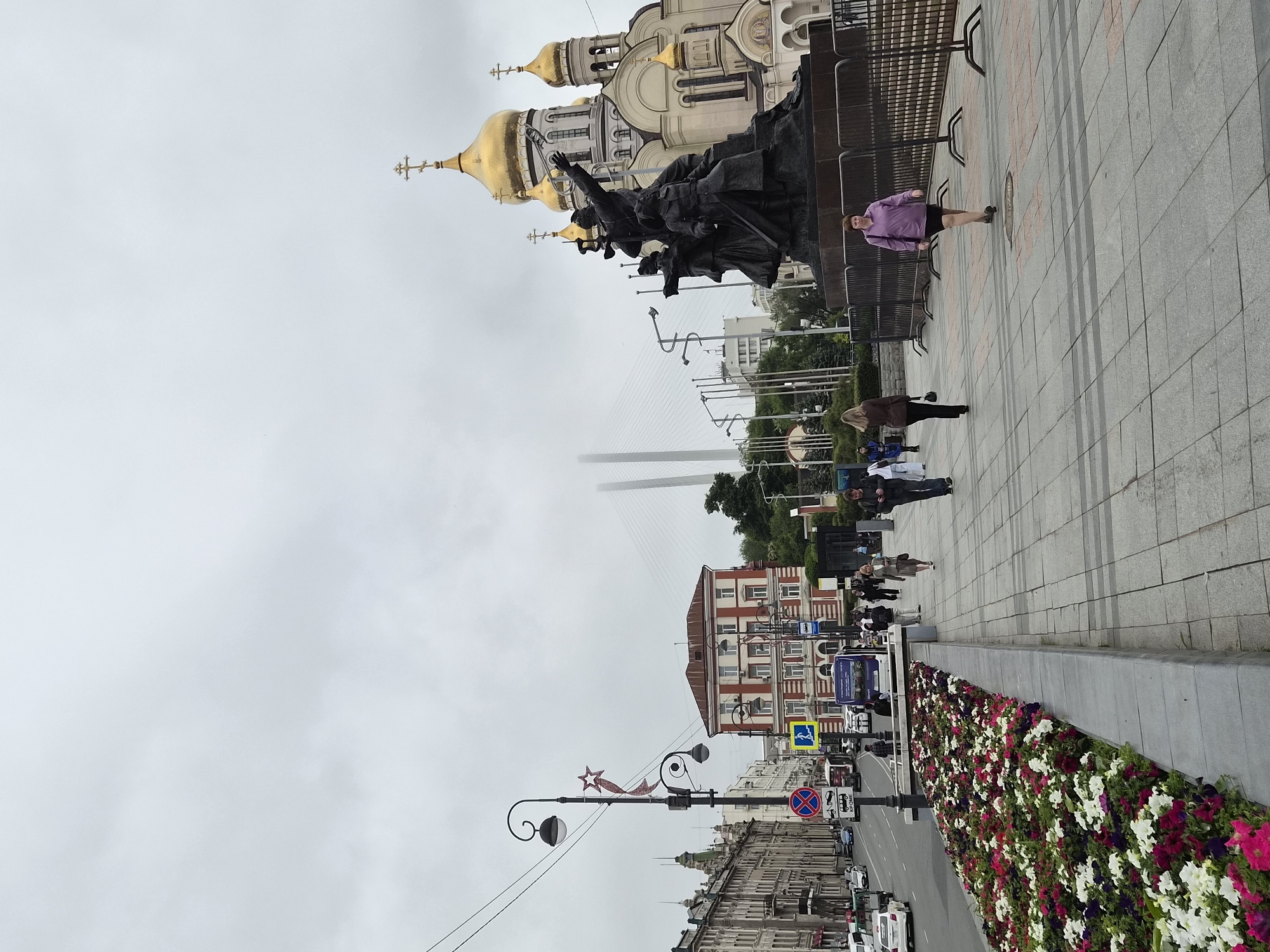 This is a full-frame, eye-level, vertical, outdoor, daytime, medium shot of a street scene in Vladivostok, Russia. The image captures the monument to the Fighters for Soviet Power in the Far East, with the golden domes of the St. Nicholas Orthodox Cathedral in the background. A large, bronze monument featuring several figures, presumably representing historical revolutionaries or soldiers, stands prominently on the right side of the frame. Behind the monument, the St. Nicholas Orthodox Cathedral, a large, ornate building with multiple golden domes and crosses, is visible. The sky is overcast and hazy, obscuring parts of the distant Golden Horn Bay Bridge.
In the foreground, a paved walkway runs alongside a raised flower bed filled with a variety of colorful flowers, including petunias in shades of pink, white, red, and purple. Several individuals are walking on the walkway. On the left side of the image, a street with traffic, including cars and a bus, is visible. Buildings line the left side of the street, exhibiting architectural styles typical of urban areas. Streetlights and traffic signs are also present. A woman in a purple shirt and dark shorts walks towards the viewer on the right side of the walkway. To her left, other pedestrians are visible, some walking away from the camera and others towards it. The overall scene depicts a public space in Vladivostok, likely a popular tourist or civic area, under cloudy weather conditions.