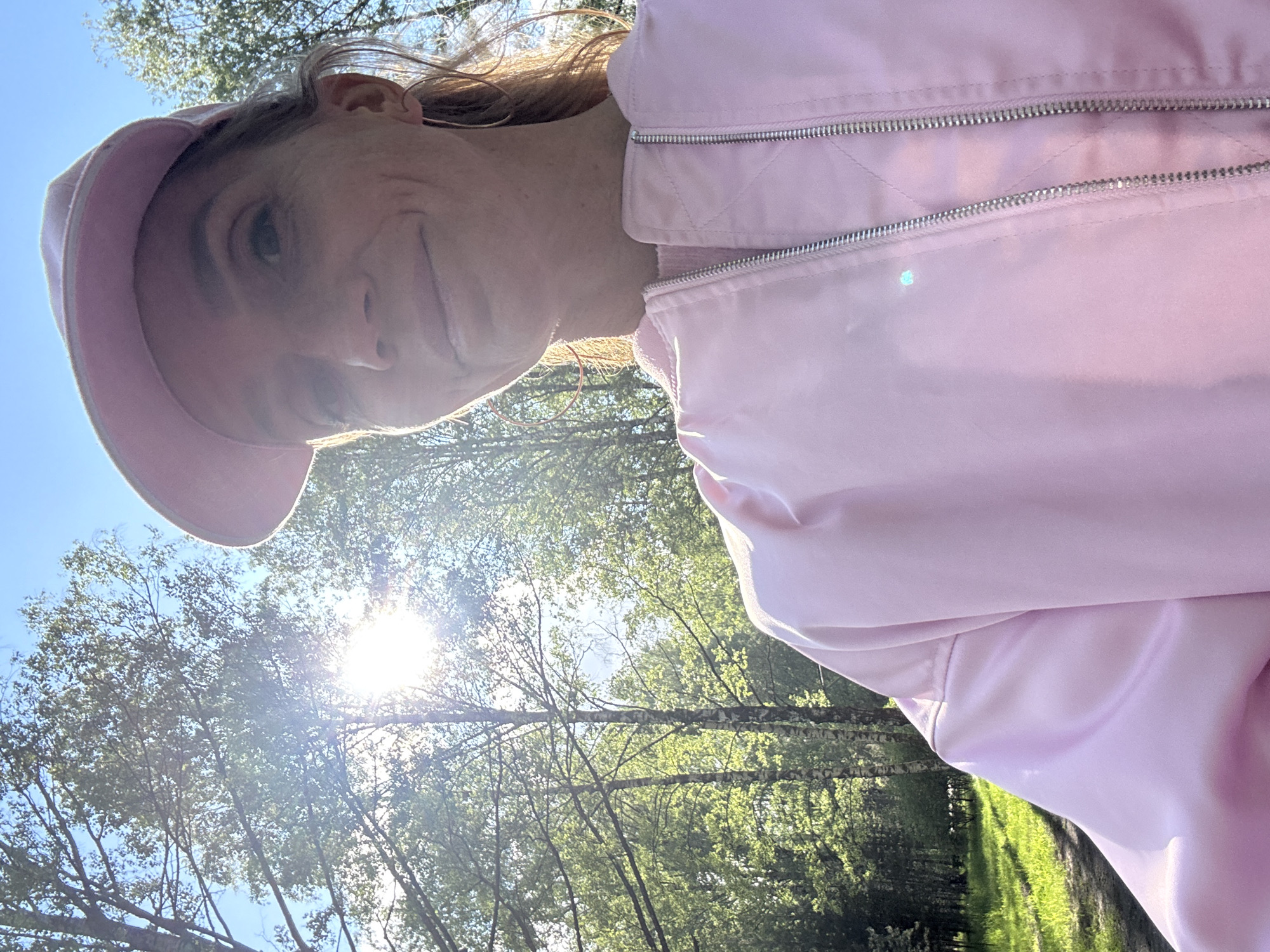 The image is taken from a low angle, looking up at a woman wearing a pink cap and a pink bomber jacket. The background shows a forest with green trees and a bright sky with the sun shining through the trees. The location is a wooded area, possibly a park or forest on the outskirts of an unknown city in Russia. The woman is looking down at the camera.