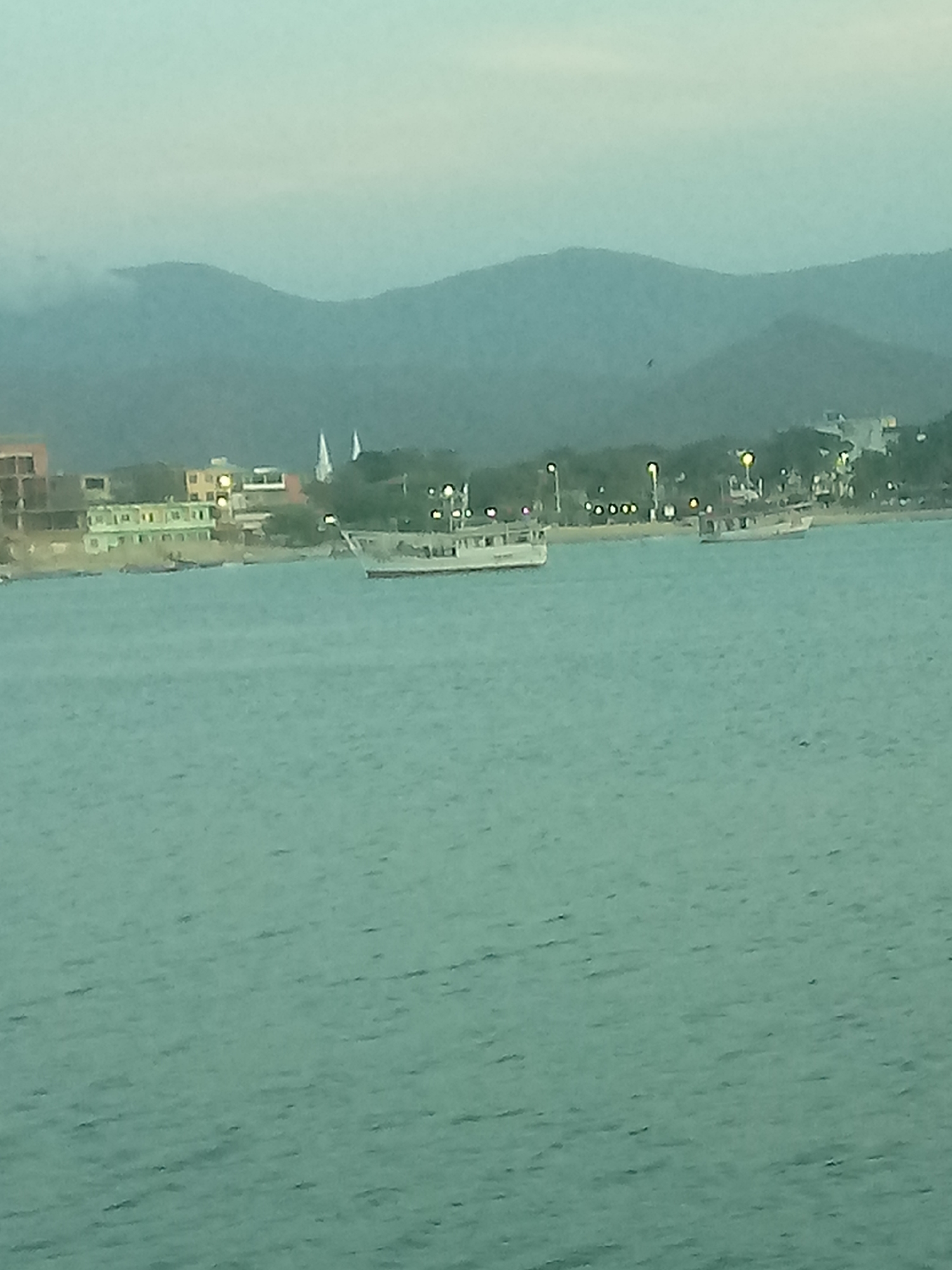 The image depicts a view of the bay in Juan Griego, Venezuela. In the foreground is the greenish-blue water of the bay. Several boats are present on the water's surface, including one larger boat in the center of the frame. The shoreline is visible in the mid-ground, lined with buildings, trees, and streetlights. Two white spires, potentially part of a church, rise above the other structures along the shore. The backdrop consists of hills and mountains in the distance, partly obscured by haze or clouds. The sky is overcast, suggesting either early morning or late afternoon light.