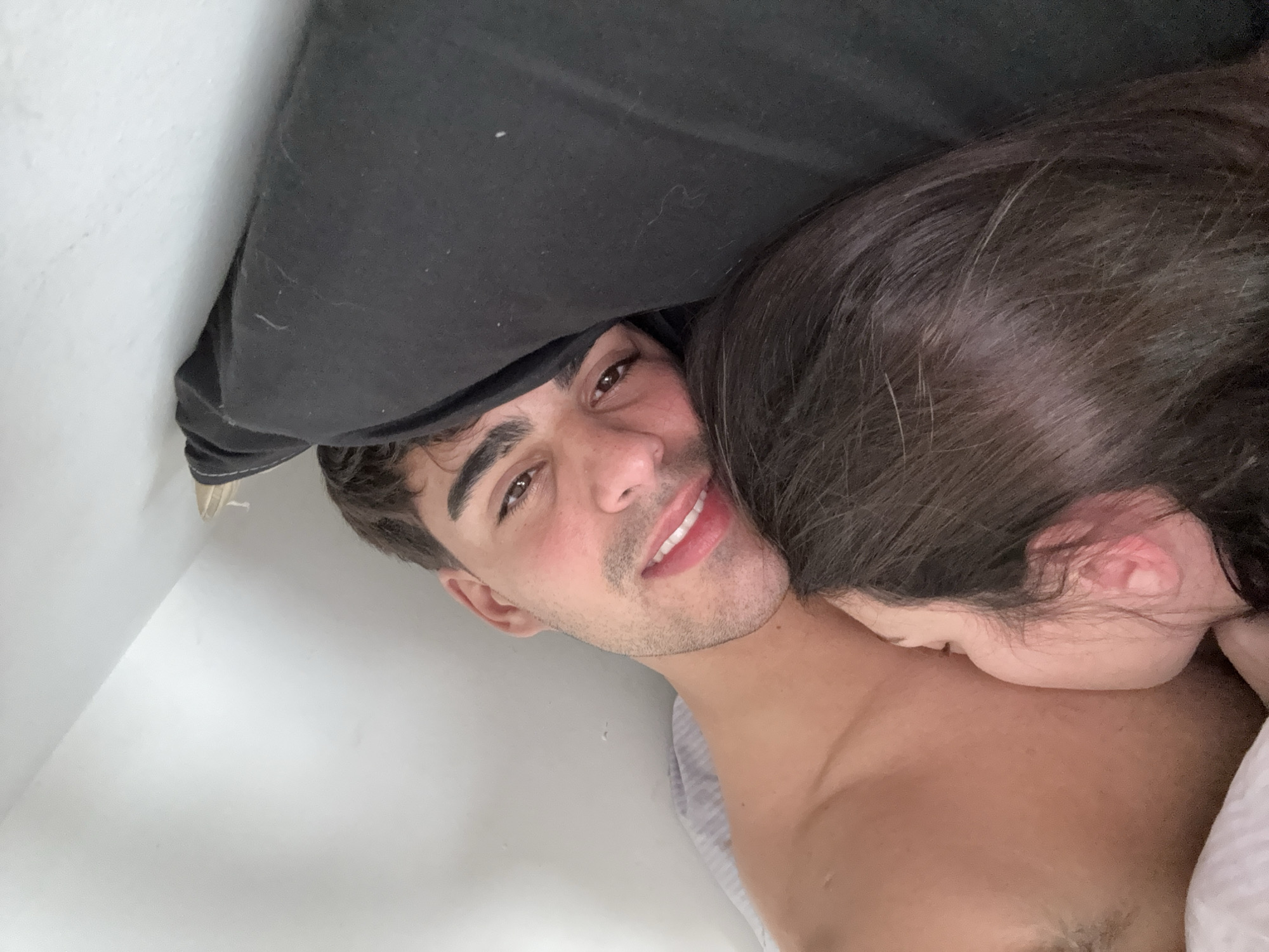 The image shows a close-up, head-and-shoulders shot of two individuals in a bed, likely in a domestic setting in Avellaneda, Argentina. One individual, appearing to be male, is facing the camera with his eyes open and a slight smile, showing his teeth. He has dark hair and eyebrows. The other individual, appearing to be female, is partially visible on the left, with dark hair resting on the shoulder of the male. Both are positioned closely together, suggesting intimacy. They are lying on a black pillow, and a white sheet is visible in the background. The lighting suggests it may be morning or a dimly lit room. There are no external environmental cues indicating a specific city location.
