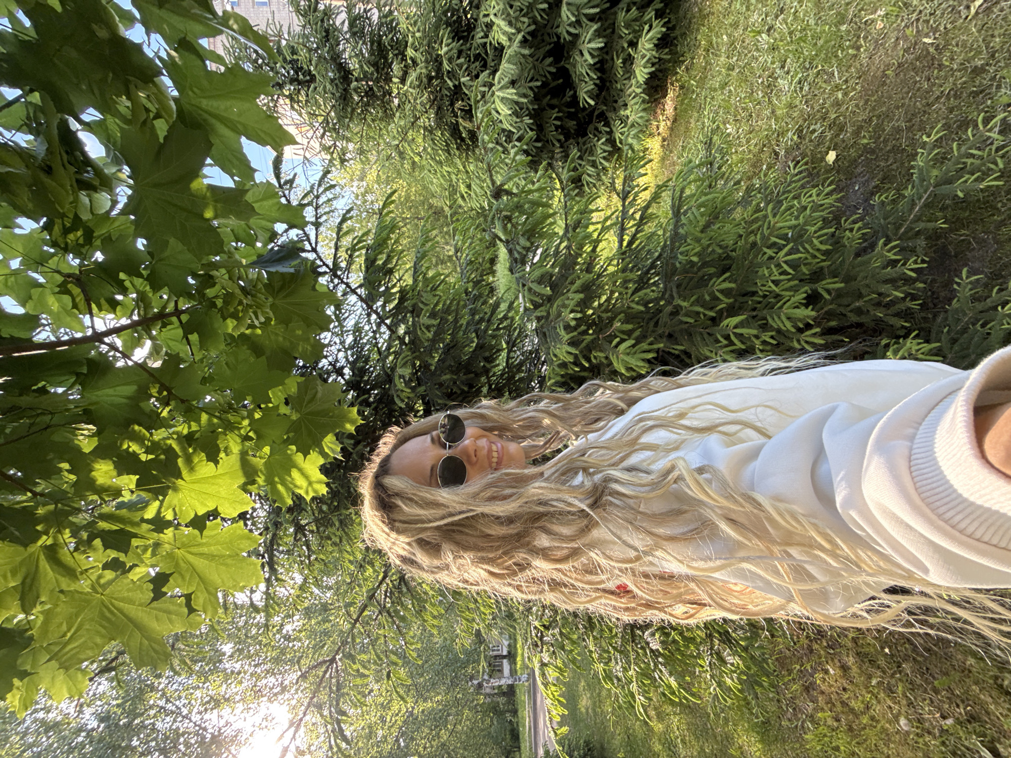 A person is featured in a bright outdoor selfie, smiling broadly with long, wavy blonde hair illuminated by direct sunlight. They are wearing round sunglasses and a light-colored (white or off-white) long-sleeved top or sweatshirt. The image is oriented sideways, giving the perspective of looking up into a vibrant green landscape.
The setting is a verdant park or natural area in Rusanovo, Russia. Large, bright green maple leaves fill the upper left foreground, providing dappled shade. The background is dense with various types of foliage, including coniferous trees with needle-like branches and other deciduous trees, some with slender, drooping leaves. Patches of green grass and ground cover are visible on the right. In the distance, a narrow path or road can be seen through the trees, and a distant building, possibly an apartment block, is faintly visible against the sky.
The strong, warm sunlight filtering through the leaves suggests either late afternoon or early morning, creating a "golden hour" effect that highlights the person's hair and adds to the cheerful atmosphere. The person's wide smile conveys happiness and relaxation. No visible text is present in the image.