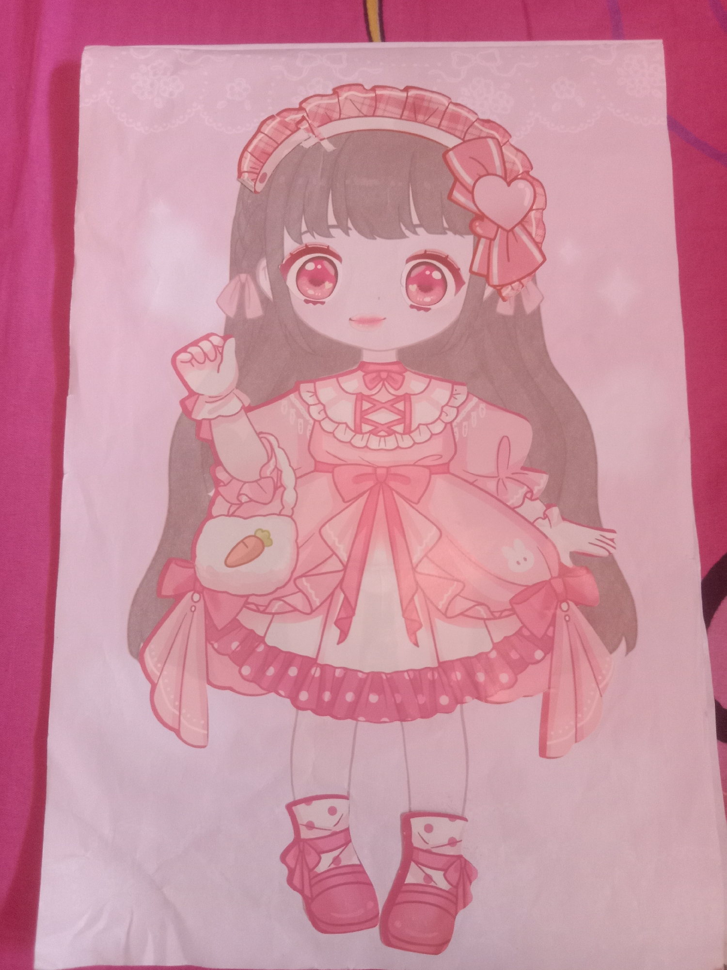 A drawing of a cartoon girl in a pink dress and matching shoes is displayed on a pink fabric surface, likely a tablecloth or bedsheet. The drawing features the girl with large, sparkling eyes, dark hair, and a headband adorned with a pink bow and heart. She is holding a small, white purse with a carrot design. The dress is layered and frilly, with a dotted hemline and bows. Her shoes are pink Mary Janes with straps. The background of the drawing has a lace-like pattern. The image is captured from a top-down perspective, and there are no visible elements to indicate a specific location in Kedungsari, Indonesia.