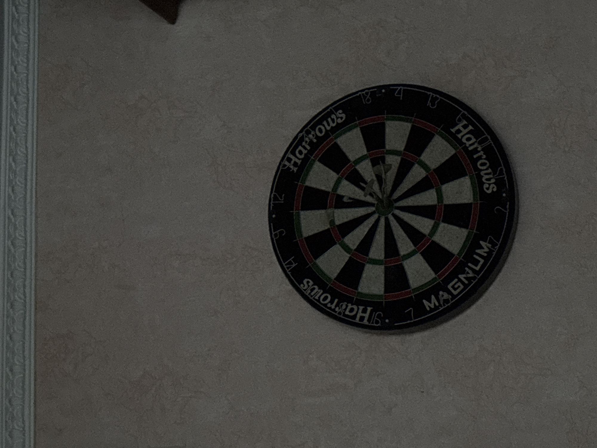 This media file shows a dartboard mounted on a textured wall. The dartboard is circular and displays the standard numbers and scoring segments of a dart game. There are several darts stuck in the board. The lighting in the image is dim, suggesting it might be evening or indoors with low light. The wall has a subtle pattern and a decorative border is visible on the left side of the frame.
Text visible on the dartboard includes "Harrows" written in a semi-circle above the board, and "MAGNUM" written in a similar fashion below. The numbers from the dartboard scoring system are also visible, such as 18, 4, 13, 2, 7, 16, and 14. The overall scene is still and quiet, with no people present. The focus is on the dartboard itself as an object within a room.