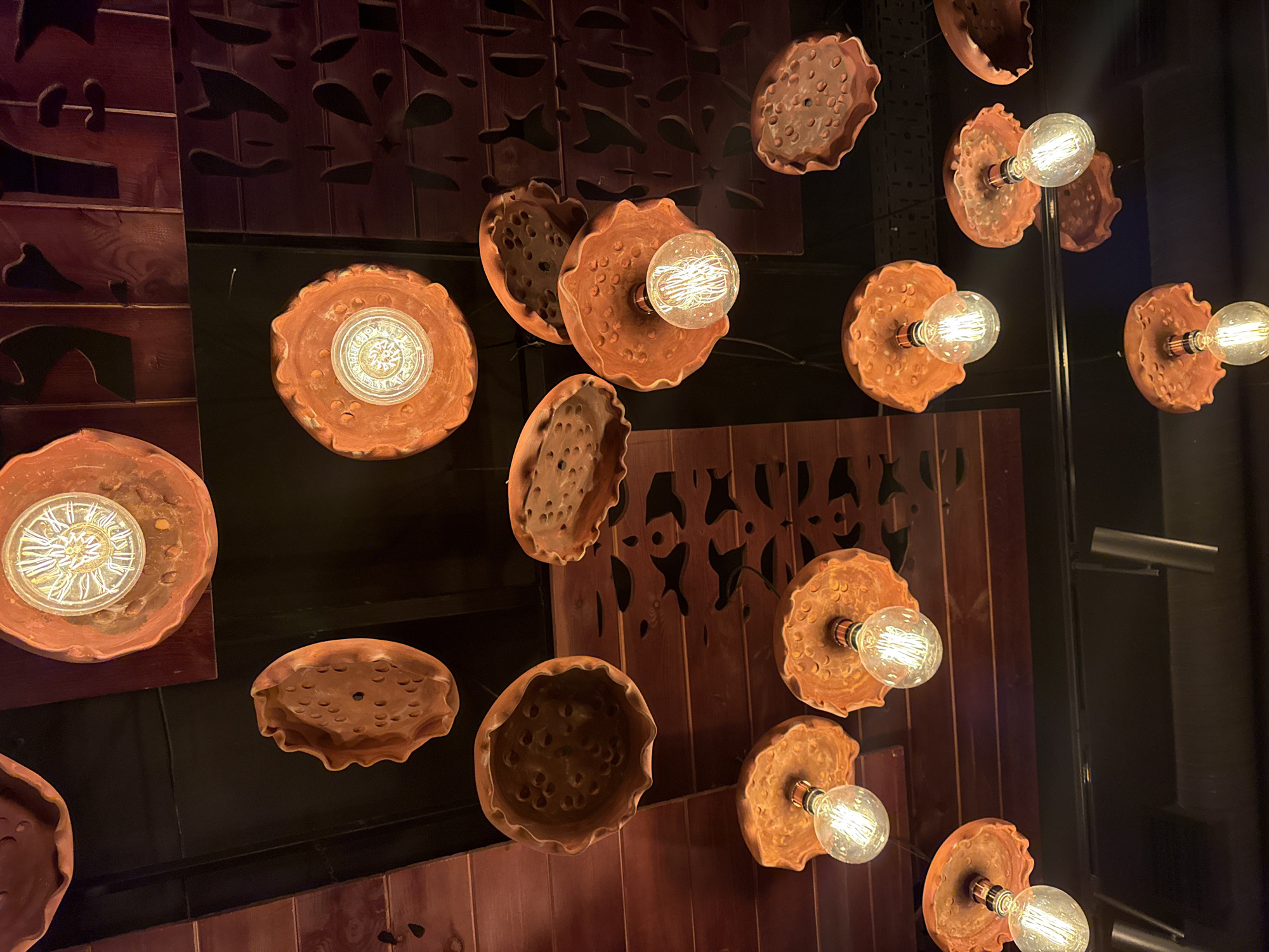 The image shows a collection of decorative ceiling lights. These lights have a reddish-brown, textured ceramic base that resembles a ruffled bowl or a porous organic shape. Each base holds a clear Edison-style light bulb with visible filaments. The lights are suspended from a dark ceiling, with wooden panels and decorative cutouts visible in the background. The lighting is warm, casting a soft glow on the objects. The scene is a well-lit interior, likely a restaurant or a stylish commercial space. There are no people visible in the image. The overall aesthetic is rustic and artistic, with an emphasis on natural materials and textured forms. The location is not specified within the image.