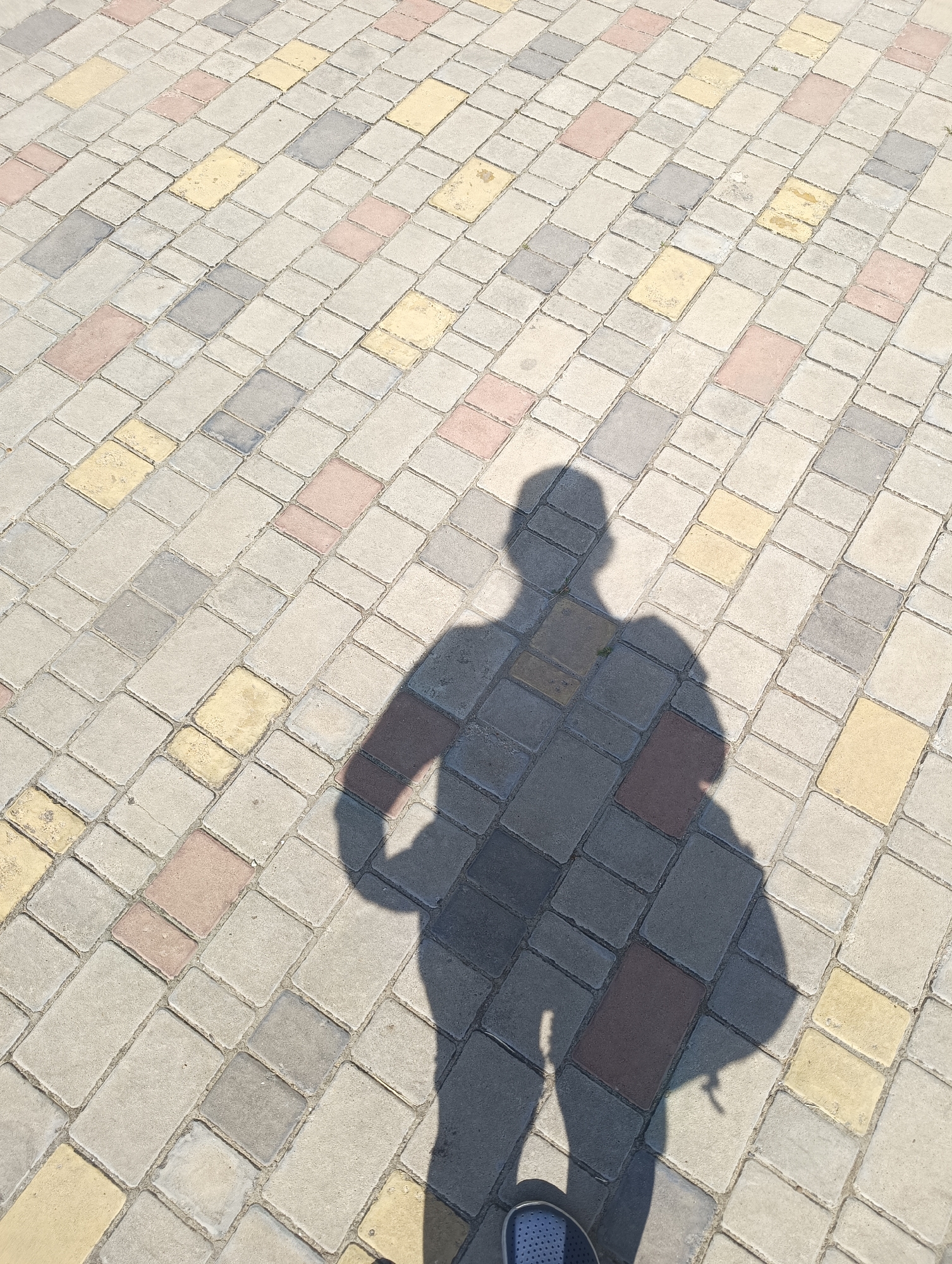 The image displays a top-down view of a paved ground surface in Maiaky, Ukraine, illuminated by direct sunlight which casts a distinct human shadow. The pavement is composed of rectangular interlocking blocks of various hues including light grey, darker grey, pale yellow, and reddish-brown, arranged in a diagonal pattern across the frame. A prominent, sharply defined shadow of a person is centered in the lower half of the image, depicting a human figure with arms bent and hands positioned on the hips. The lower-left corner of the frame shows the toe area of a blue shoe with a white sole, confirming the presence of the individual casting the shadow and taking the photograph. No other subjects or objects are discernible within the visible area.