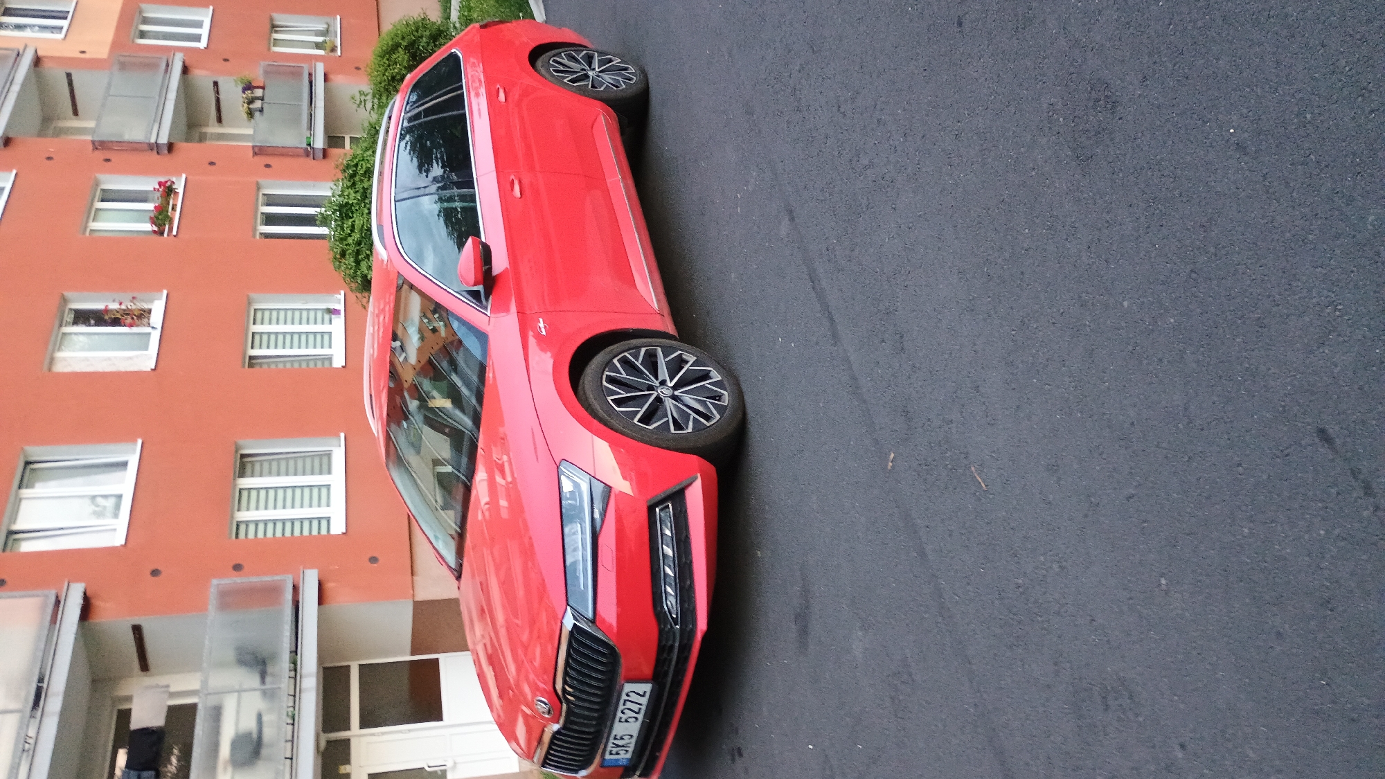 A red Skoda Superb estate car is parked on an asphalt surface in front of a multi-story residential building with orange-colored facade. The car's license plate reads "5K5 5272". The building has multiple windows with white frames and blinds, some with small flower boxes. A balcony is visible on the left side of the building with some laundry hanging. Green bushes are present near the building. The scene appears to be outdoors during daylight.