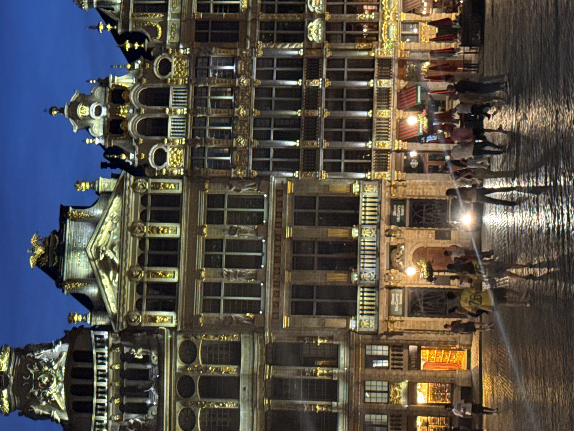 This is a medium-distance, eye-level night shot of the Grand Place in Brussels, Belgium. The ornate historical buildings of the square are illuminated, showcasing their intricate architectural details and golden embellishments. The sky above is a deep twilight blue. The cobblestone ground is wet and reflects the lights of the buildings and streetlamps, creating a shimmering effect.
In the foreground, a group of approximately 15 people are scattered across the square. They appear to be tourists or locals enjoying the evening ambiance. Some are walking, others are standing and talking. A few individuals are wearing bright clothing, such as a person in a yellow shirt with the number "26" visible. A car with its headlights on is also present, suggesting it is driving through the square.
The buildings feature multiple stories with numerous windows, balconies, and decorative facades. Some ground-level establishments have their entrances open, with interior lights illuminating the spaces. A few of these businesses have awnings visible. The overall scene conveys a lively yet serene atmosphere characteristic of a European city square at dusk.