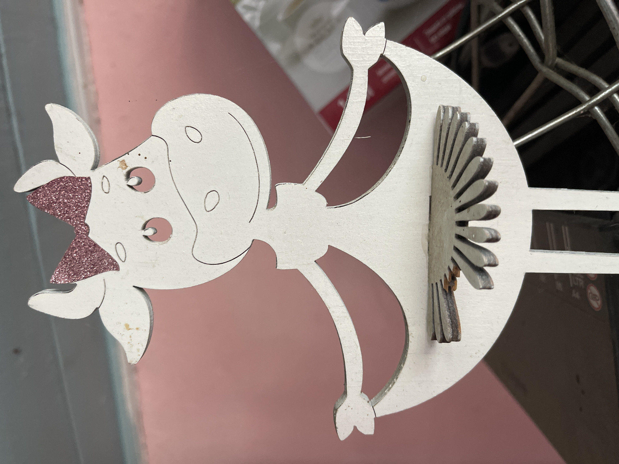A close-up, eye-level, slightly angled shot features a white, wooden cutout of a cartoon cow. The cow is depicted from the chest up, wearing a pink glitter bow on its head. It has large, pink circular eyes with black pupils and a small black dot above its right eye. Two small, curved black lines represent nostrils, and a wider black line forms its smile. The cow's body transitions into a flared skirt-like shape, with stylized arms extended outwards. A decorative, fan-shaped element made of gray metal with etched lines is attached to the inside of the skirt. The background is divided, with the top section showing a dusty, light blue surface, and the bottom section a soft pink. The overall scene is indoors, with a hint of a wire rack and some blurred objects in the lower right corner. There is no indication of Aleksandrovskoye, Russia, or any specific location.