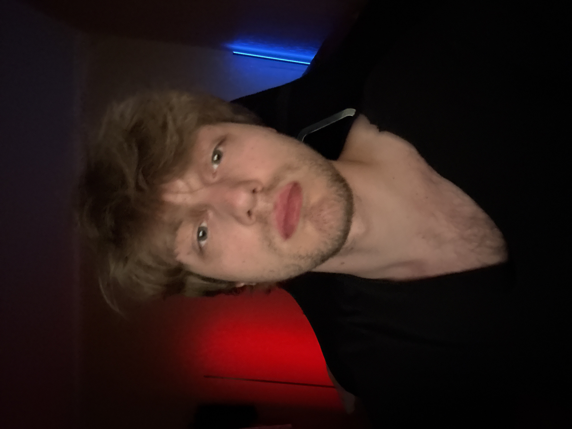 The image is a close-up, sideways portrait of a young Caucasian man. He has light brown, messy hair and appears to be looking slightly off-camera with a neutral to slightly confused expression. His eyes are light blue. He has stubble on his face. He is wearing a black shirt, the collar of which is visible. Behind him, there is a red light illuminating the lower portion of the scene and a blue light strip on the upper wall. The lighting creates a moody and intimate atmosphere. The background is dark, suggesting the photo was taken indoors, possibly at night or in a dimly lit room. There are no visible objects or text in the image. The setting appears to be a personal space, perhaps a bedroom or living area, given the ambient lighting. The overall impression is of a candid shot, possibly a selfie, taken in a relaxed and private environment.