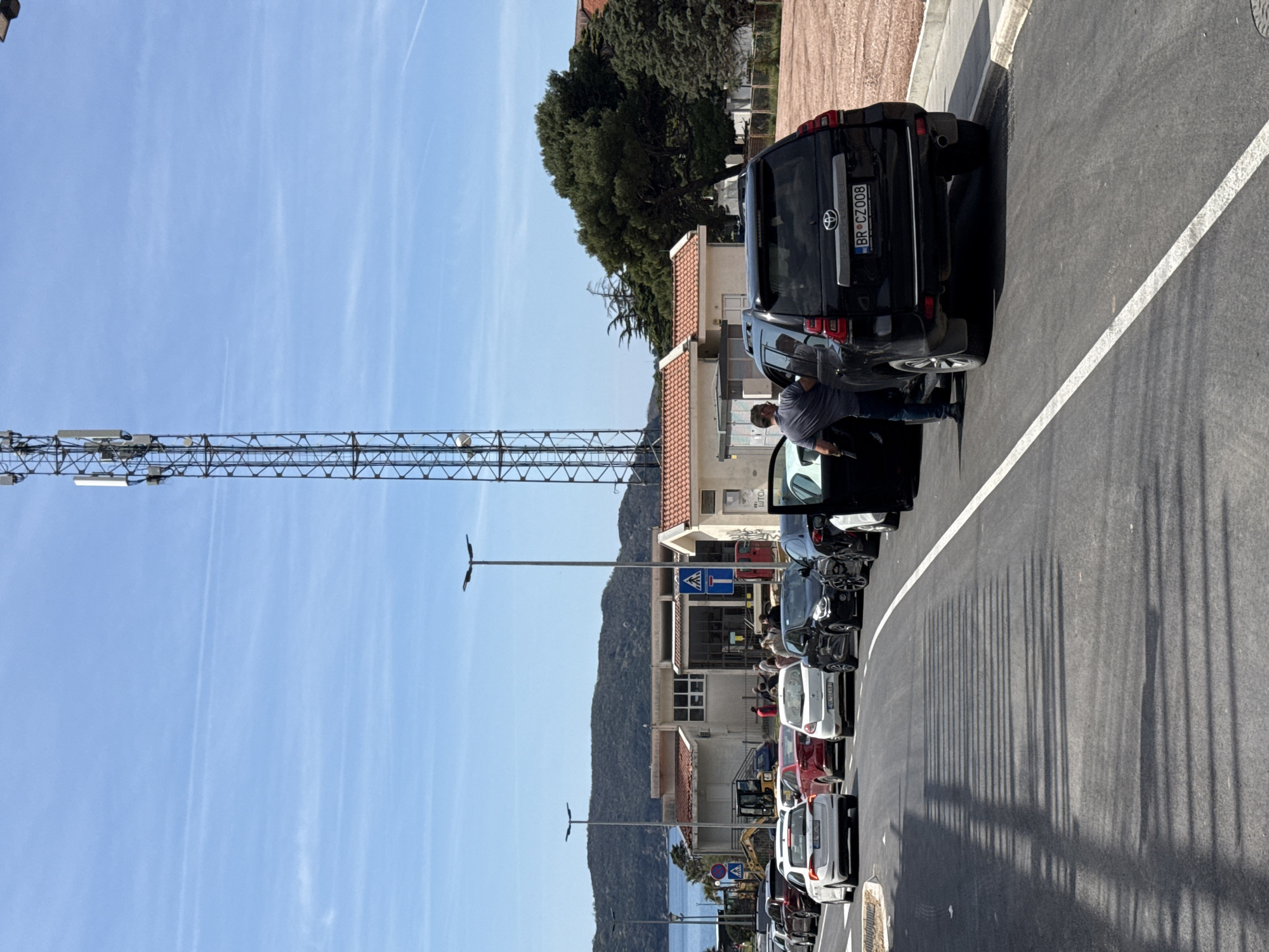 A street in an unknown city on an unknown date. A row of parked vehicles lines the street, with a person standing next to a black SUV. The vehicle's license plate reads "BR CZ008". In the background, buildings and a mountain can be seen, and a communications tower is visible in the sky. The weather is clear, with a blue sky and some clouds. The scene is captured during daylight, and long shadows are cast by the streetlights onto the asphalt.