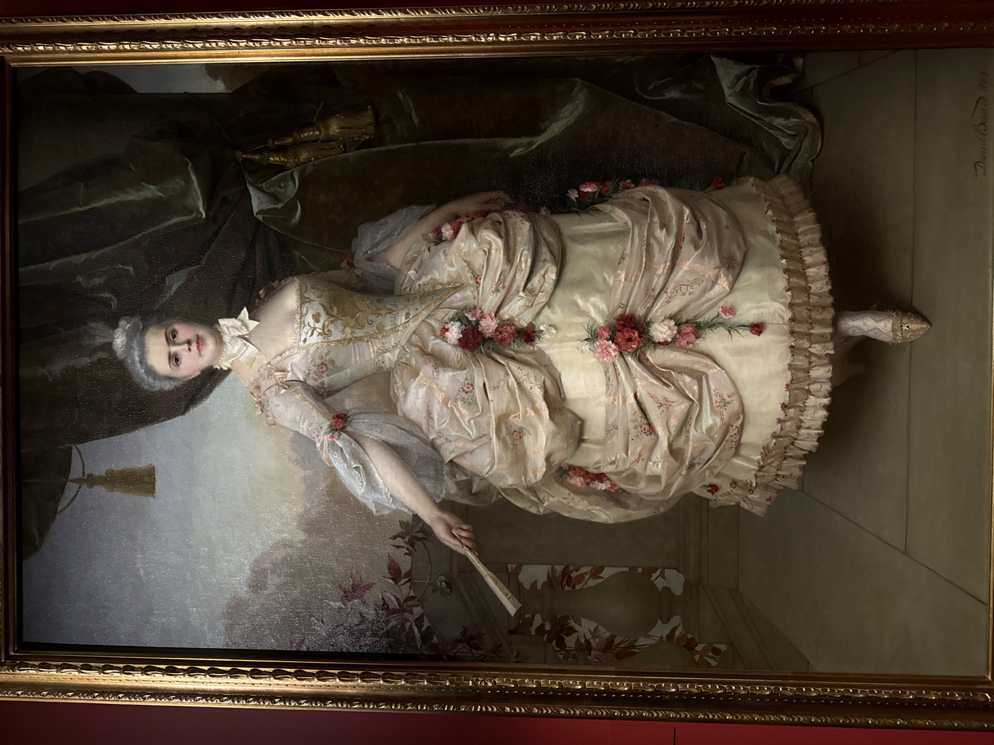 The image is a framed portrait of Madame Récamier, painted by Jacques-Louis David in 1800. Madame Récamier reclines on a chaise lounge in a neoclassical interior. She is dressed in an empire-waisted gown of ivory and cream, adorned with pink floral embellishments. Her hair is styled in a simple updo, and she holds a hand fan. A dark green drapery is in the background. The painting hangs in the Louvre Museum in Paris, France.