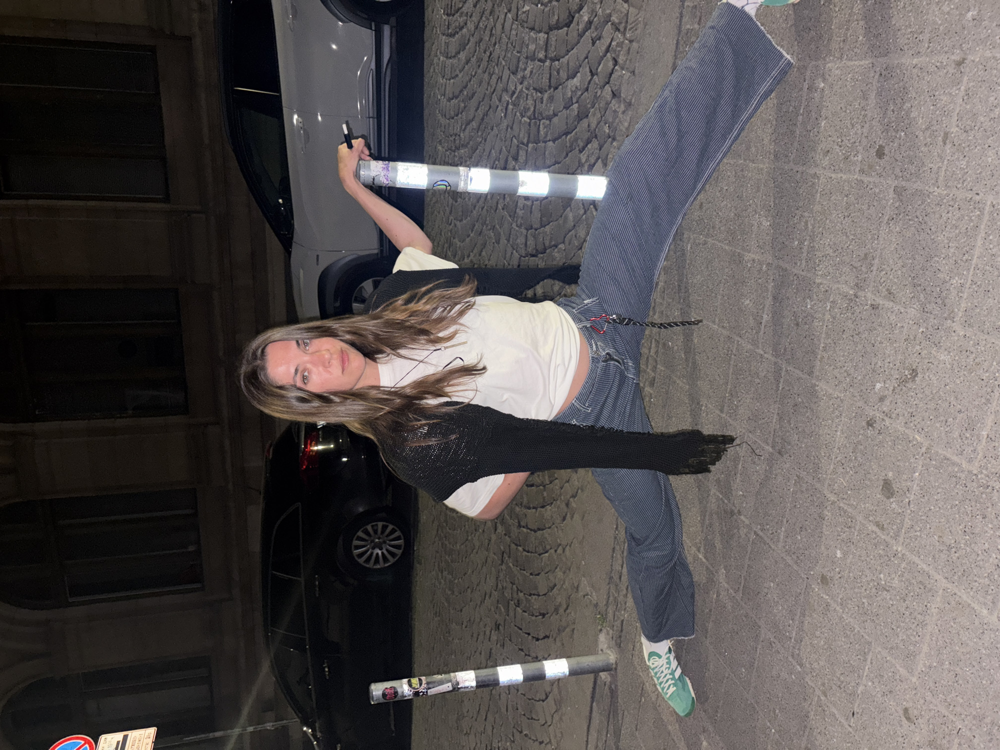 A young adult female is captured in a full-length, eye-level shot, performing a straddle split on a cobblestone street in Sofia, Bulgaria, at night. She has long brown hair and is wearing a white t-shirt, a black knit shrug, vertically striped wide-leg pants, and green and white Adidas sneakers on her left foot. Her left arm is bent behind her lower back, and her right hand rests on the top of a light grey cylindrical street bollard, while holding a slim, dark object. She maintains direct eye contact with the camera with a neutral expression. To her immediate left, a similar dark grey street bollard with white reflective strips and various stickers is visible. The foreground consists of square concrete pavers, transitioning to dark grey cobblestones where the woman is positioned. In the background, a dark-colored SUV and a light-colored SUV are parked on the street. Behind the vehicles, dark buildings with arched windows are discernible. On the far left, a red circular "No Entry" sign and a white rectangular sign with Cyrillic text are mounted on a pole.