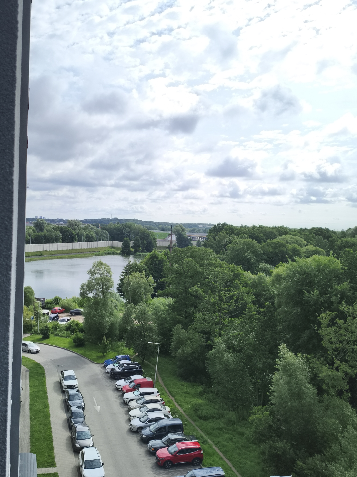 An aerial view captures a residential area bordering a serene lake, with lush green trees dominating the landscape. The sky is partly cloudy, with fluffy white clouds interspersed with patches of blue. In the foreground, a building's exterior wall, featuring a textured gray surface with a white vertical line, is visible on the left. A paved road curves through the scene, lined with parked cars of various colors and models. A white arrow painted on the road indicates a one-way direction. Beyond the road, a body of water reflects the overcast sky. A concrete barrier separates the water from a dense forest, which stretches towards the horizon. Distant buildings and structures can be seen peeking through the trees, suggesting a semi-urban environment. No people are visible in the frame, and no particular actions or interactions are taking place. The overall impression is one of a peaceful, nature-infused urban setting. The presence of modern buildings and infrastructure hints at a developed area within a city. Given the context of Kaliningrad, Russia, this scene likely depicts a residential development with a recreational waterfront area.