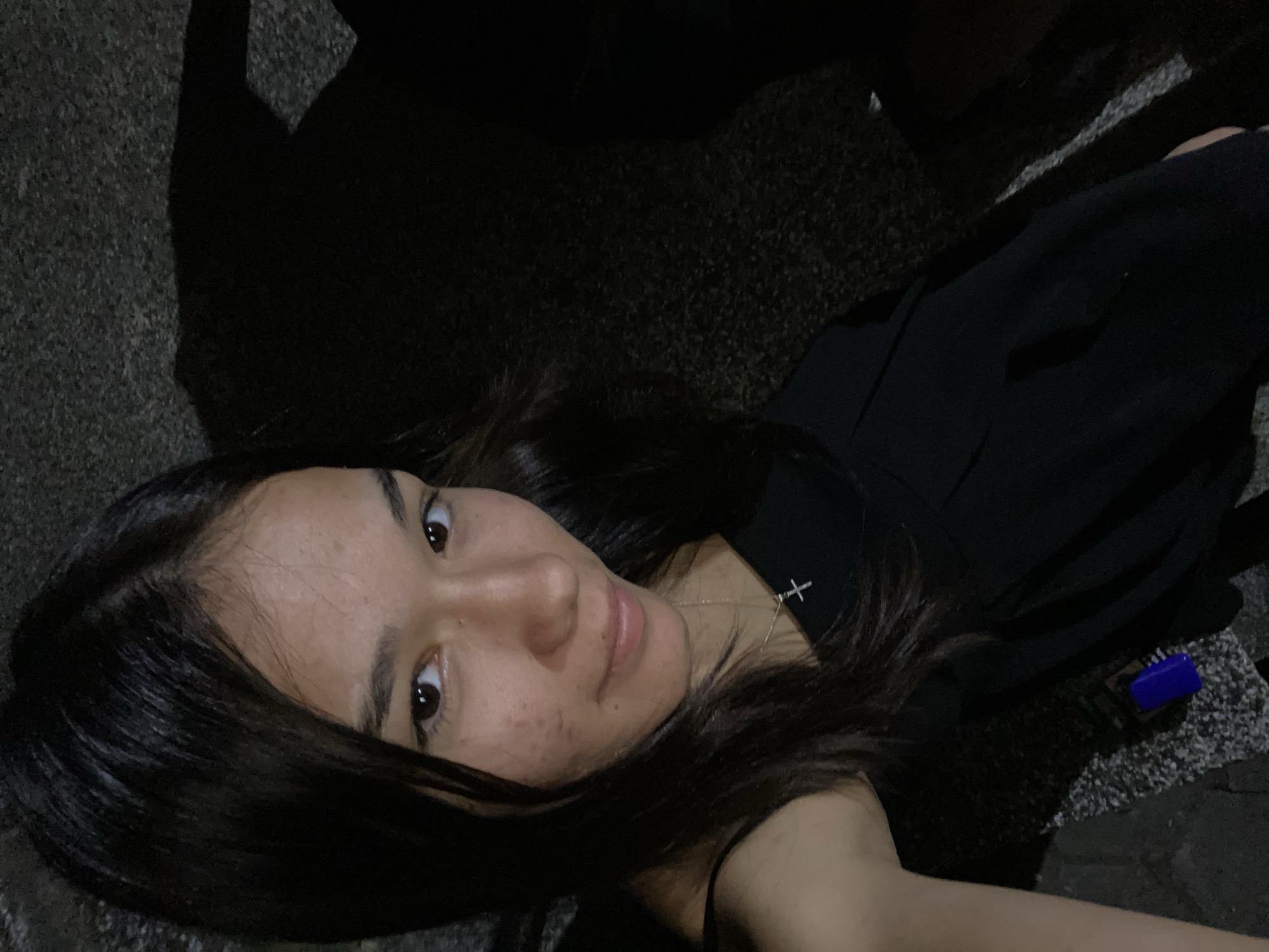 A young woman with dark hair and brown eyes is taking a selfie. She is wearing a black dress and a silver cross necklace. She is standing outdoors at night, on what appears to be a street or sidewalk. The background is dark, with shadows visible. There is a blue object on the ground near her. The scene does not contain any identifiable landmarks or characteristics that would place it specifically in Hirevo, Bulgaria.