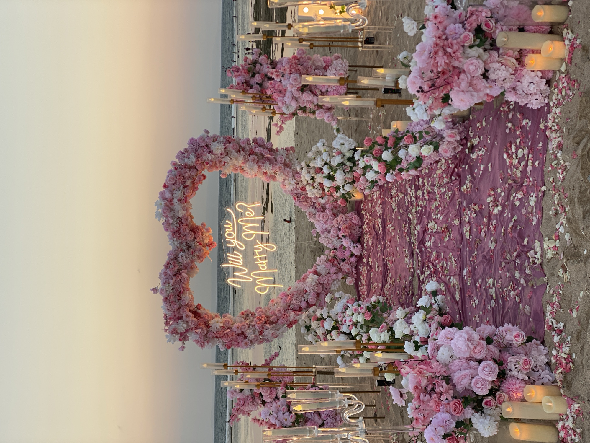 A romantic marriage proposal setup is arranged on a beach in Kuta Selatan, Indonesia, at dusk. A large heart-shaped arch, densely adorned with pink and white flowers, frames the ocean and sky. Within the arch, a neon sign spells out "Will you Marry Me?" in a cursive font. A pathway covered with a dark pink cloth and scattered flower petals leads to the arch. Flanking the pathway are tall candelabras with faux candles, and large floral arrangements in shades of pink and white. Several real candles are also placed on the sand. In the background, people can be seen walking along the shoreline, and a lone individual is seated in the shallow water. The sky displays a soft gradient of orange and yellow, indicative of a sunset.