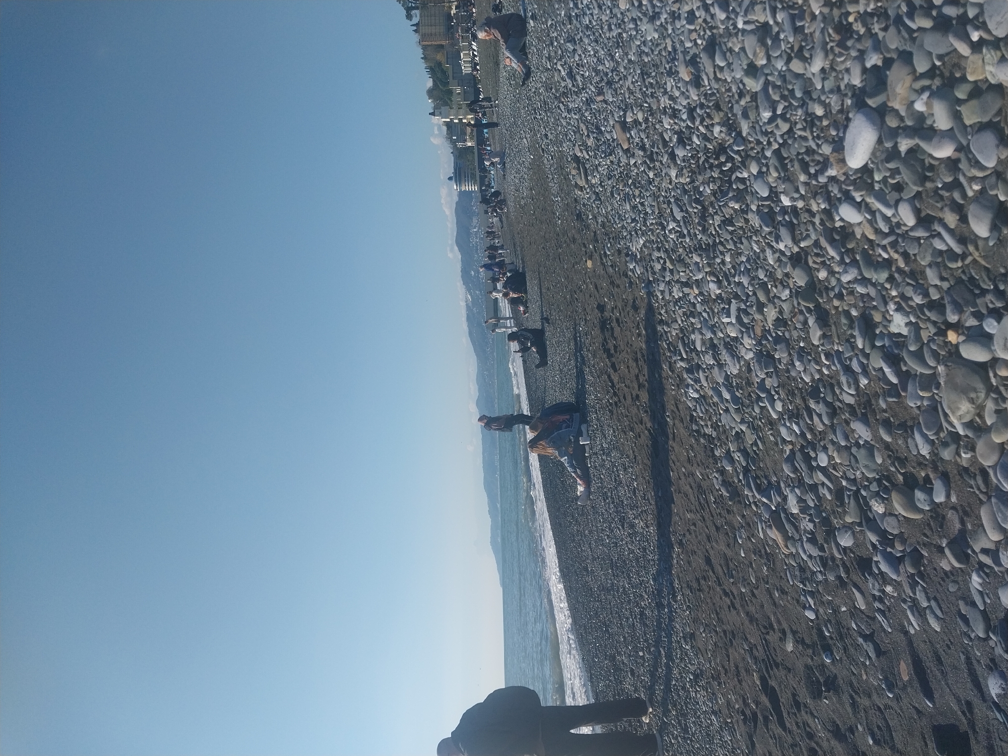 This image captures a wide shot of a pebble beach in Sochi, Russia, under a clear, bright blue sky. The picture is oriented vertically, showing the sky on the left and the beach and sea on the right.
The scene depicts a calm day at the seaside. The left portion of the image is dominated by an expanse of unblemished, light blue sky. A clear horizon line runs diagonally across the frame, separating the sky from the coastal landscape.
The right side of the image features a expansive pebble beach, stretching towards the distant coastline. The beach consists of numerous grey and white pebbles, interspersed with darker patches of sand, particularly near the waterline. The sea, visible on the lower right, exhibits gentle, frothy waves lapping against the shore, indicating calm or moderately calm waters.
Several people are scattered along the beach. Some individuals are seen standing or walking near the water's edge, while others appear to be seated or lounging further up the pebbled expanse. Their postures suggest a relaxed and leisurely atmosphere, perhaps enjoying a stroll or soaking in the sun. Long, distinct shadows cast by the people confirm bright, clear daylight, likely mid-morning or afternoon.
In the background, along the distant horizon, a coastal cityscape with various buildings can be faintly seen, backed by a range of mountains fading into the distant haze. There is no visible text in the image. The overall impression is that of a serene and recreational coastal environment.