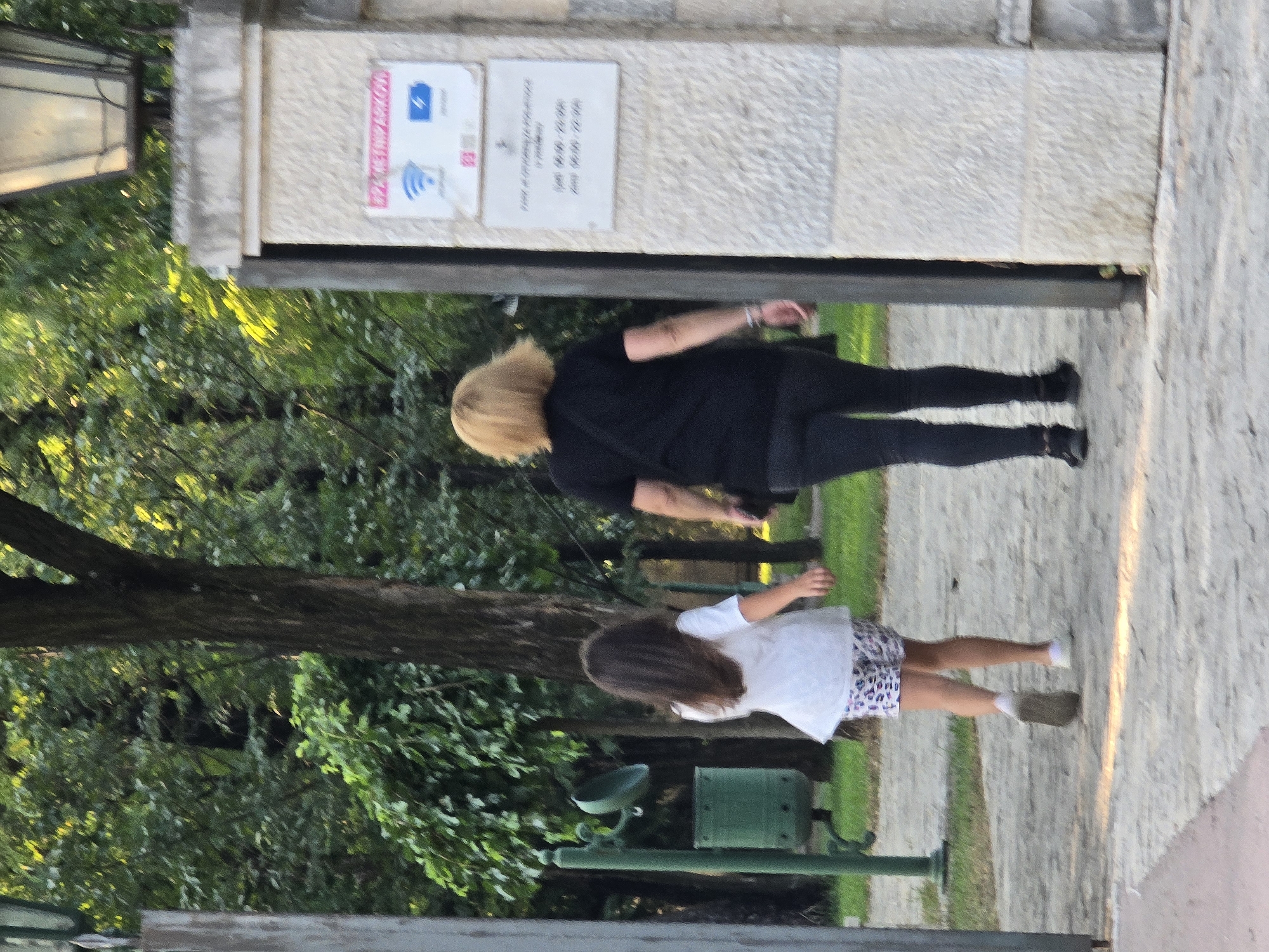 Two individuals, a young girl and a woman, walk away from the camera into a park area. The girl, on the left, wears a white t-shirt and patterned shorts. The woman, on the right, has blonde hair and wears a black t-shirt and black leggings. They are walking on a paved path with a stone pillar on the right side of the frame. A sign with symbols for Wi-Fi and a charging station is attached to the pillar. In the background, trees and foliage indicate a natural, outdoor setting. The scene appears to be an entrance to a park or public space in Podgorica, Montenegro, likely within the vicinity of a metropolitan park like Kruševac.