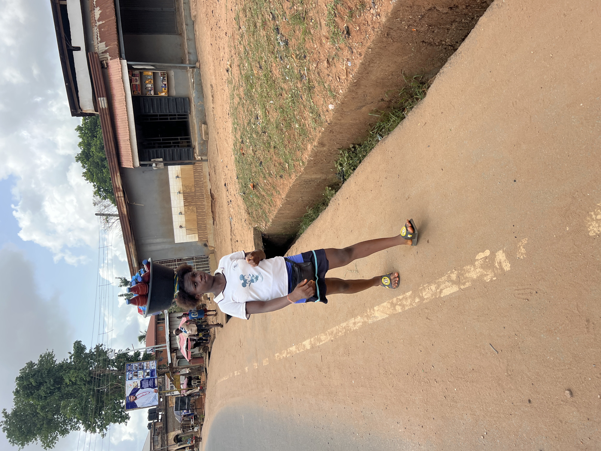 The image depicts a young woman standing on a street in Obosi, Nigeria. She is wearing a white t-shirt, blue shorts, and sandals, and she has a black fanny pack around her waist. She is carrying a large black bucket on her head, which appears to be filled with red and blue plastic bottles. The street is paved and stretches into the distance, with buildings and trees visible on either side. Other people and vehicles are present in the background. The scene appears to be a normal day in a residential or commercial area of Obosi.