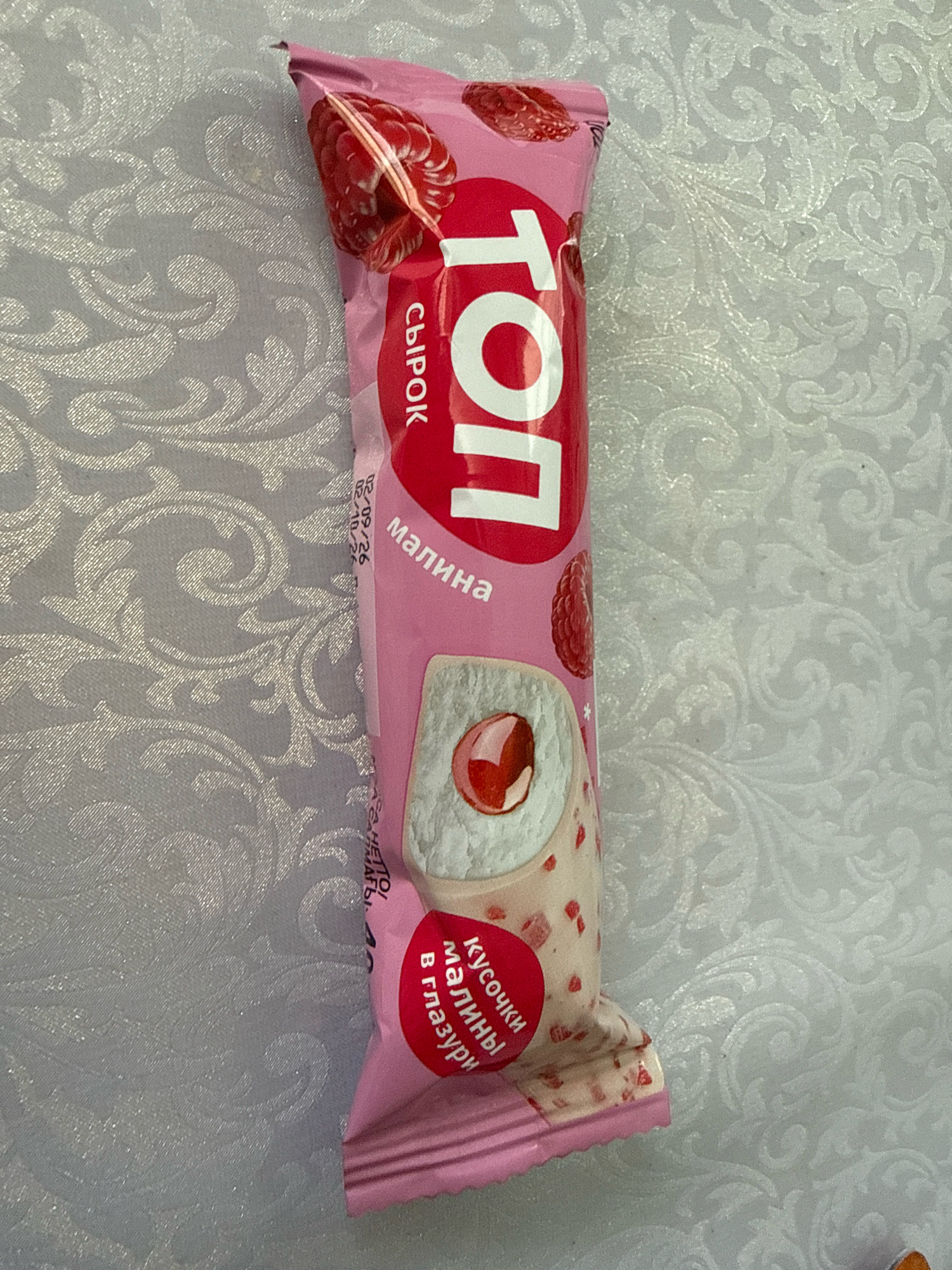 A vertically oriented, close-up shot shows a pink ice cream wrapper with a raspberry design. The wrapper features Cyrillic text, with the prominent word "ТОП" in large white letters. There is an image of a raspberry near the top of the wrapper. The ice cream itself is depicted with a white coating, studded with small red pieces and a swirl of red filling visible in a cross-section. Text on the wrapper also mentions "малина" (raspberry) and "Кусочки малины в глазури" (raspberry pieces in glaze). There is a date stamp visible on the wrapper. The background is a light-colored textured fabric. This image was taken in Kamyanyets, Belarus.