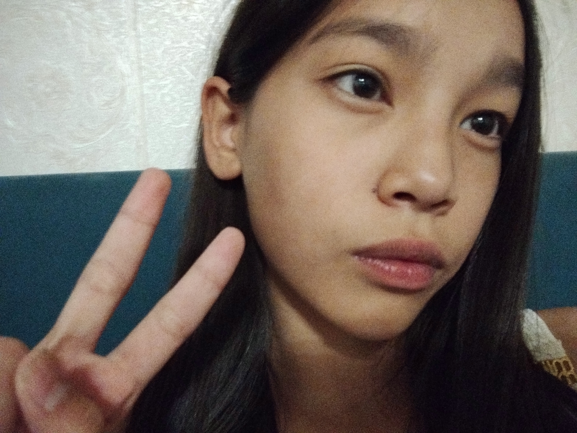 A young female with dark hair is shown in a close-up, selfie-style portrait. She is making a peace sign with her left hand in the foreground, with two fingers extended. Her face is turned slightly towards the camera, and her eyes are looking forward. She has dark eyebrows, dark eyes, and pink lips. Her skin tone appears to be light to medium. She is wearing a white and yellow patterned garment on her right shoulder. The background consists of a textured, light-colored wall and a dark blue surface behind her. The lighting suggests an indoor environment. There are no discernible landmarks or environmental cues that would allow for the precise location in Bishkek City, Kyrgyzstan to be determined from the image alone.