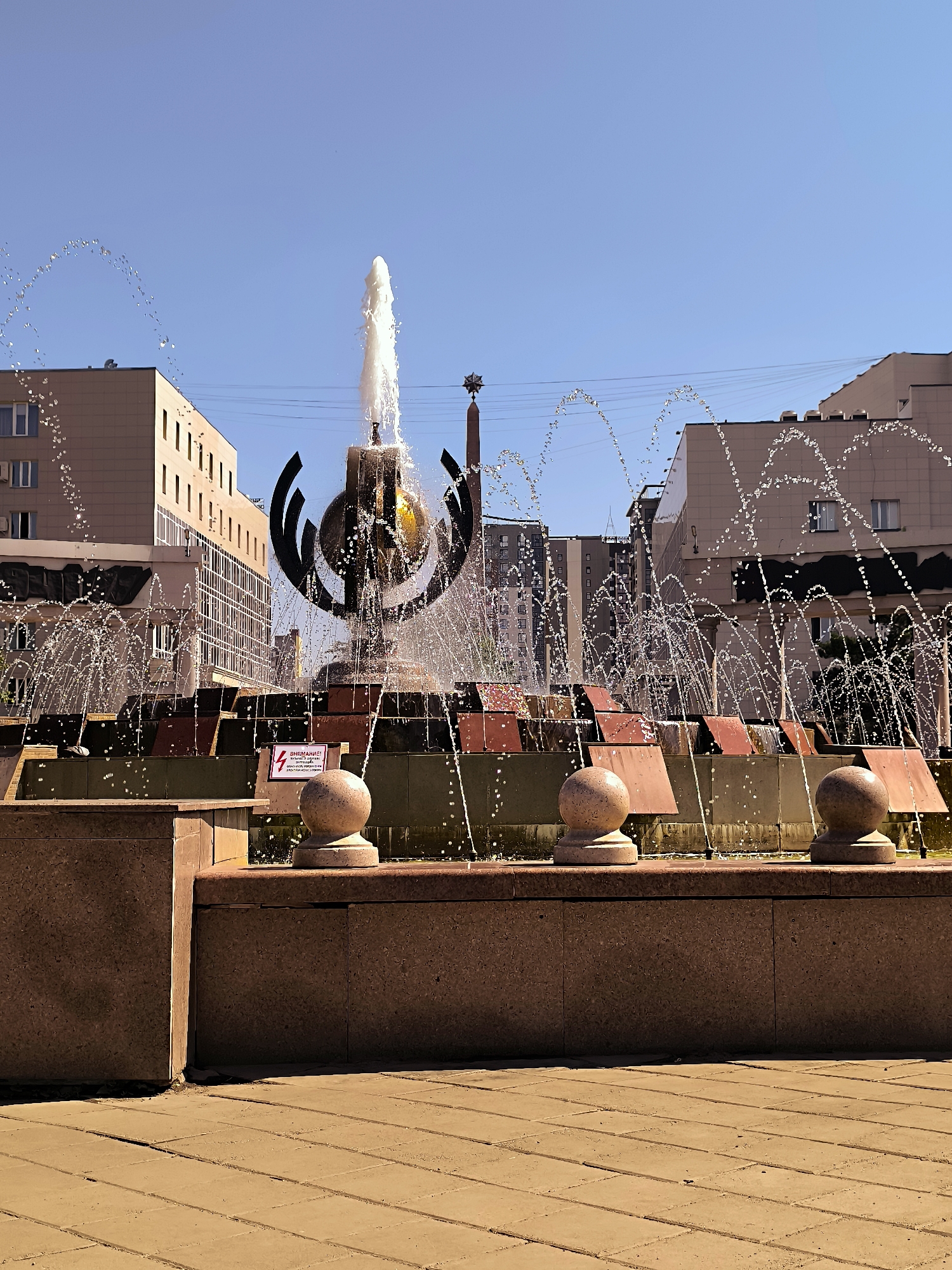 The image captures a fountain in front of the Qarağandı city administration building in Kazakhstan. The fountain features a central sculpture with water jets spraying outwards, creating arcs of water against a clear blue sky. The sculpture is dark and abstract, with curved elements surrounding a globe-like sphere. The fountain is built with tiered stone structures, and decorative stone spheres are placed on the edges of the basin. In the background, modern buildings with multiple windows line the street, and a tall obelisk with a star ornament is visible to the right of the fountain. The ground in the foreground is paved with tan interlocking blocks. There are no people visible in the image. A red and white warning sign with an electrical symbol is affixed to the fountain's structure.
