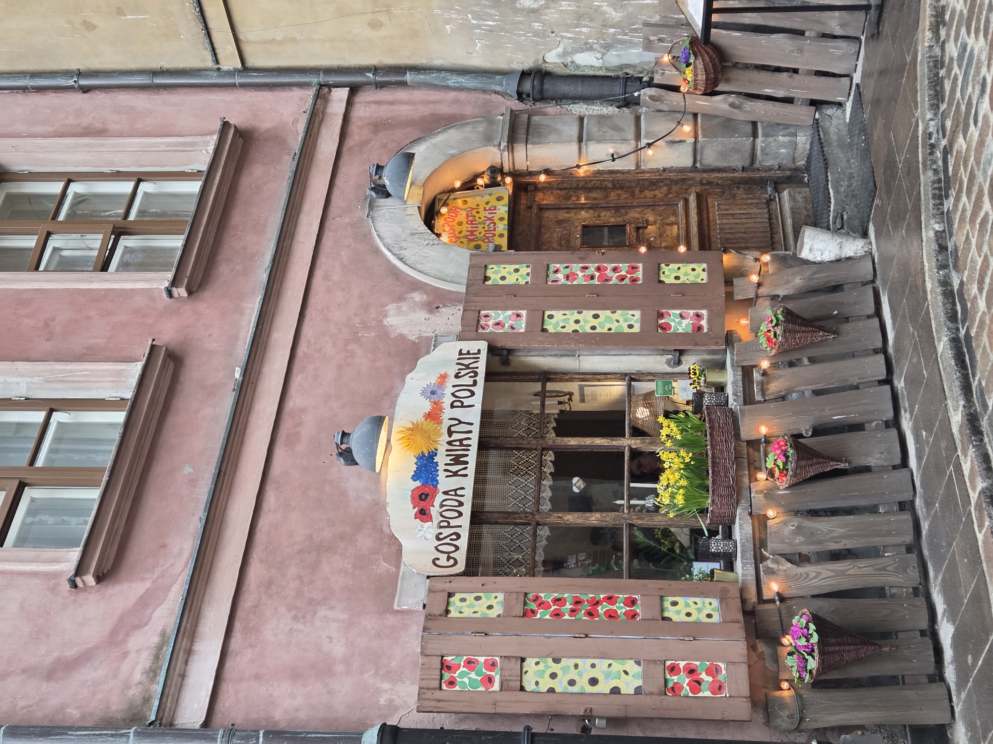 The image depicts the facade of a building in Warsaw, Poland. A pale pink building features several windows with brown frames. A shop named "Gospoda Kwiaty Polskie" (Polish Flower Inn) has a wooden sign with painted flowers. It is decorated with flower baskets, wooden planks, and fairy lights. The architecture is old, with weathered walls and a traditional design. A narrow, brick-paved alley is visible on the right side of the frame.