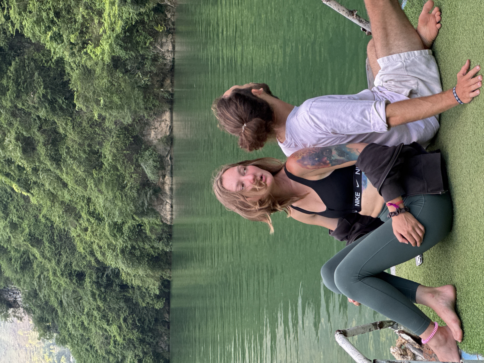 Two individuals are situated on a grassy platform, presumably a dock or similar structure, with a body of water behind them. Lush green vegetation lines the opposing bank. The woman, facing the camera, has blonde hair and wears a black sports bra with "NIKE" visible on the band, green leggings, and a dark jacket. The man, seen from the back, has long brown hair tied up and wears a white t-shirt and beige shorts. The location is Đồng Văn Commune, Vietnam.
