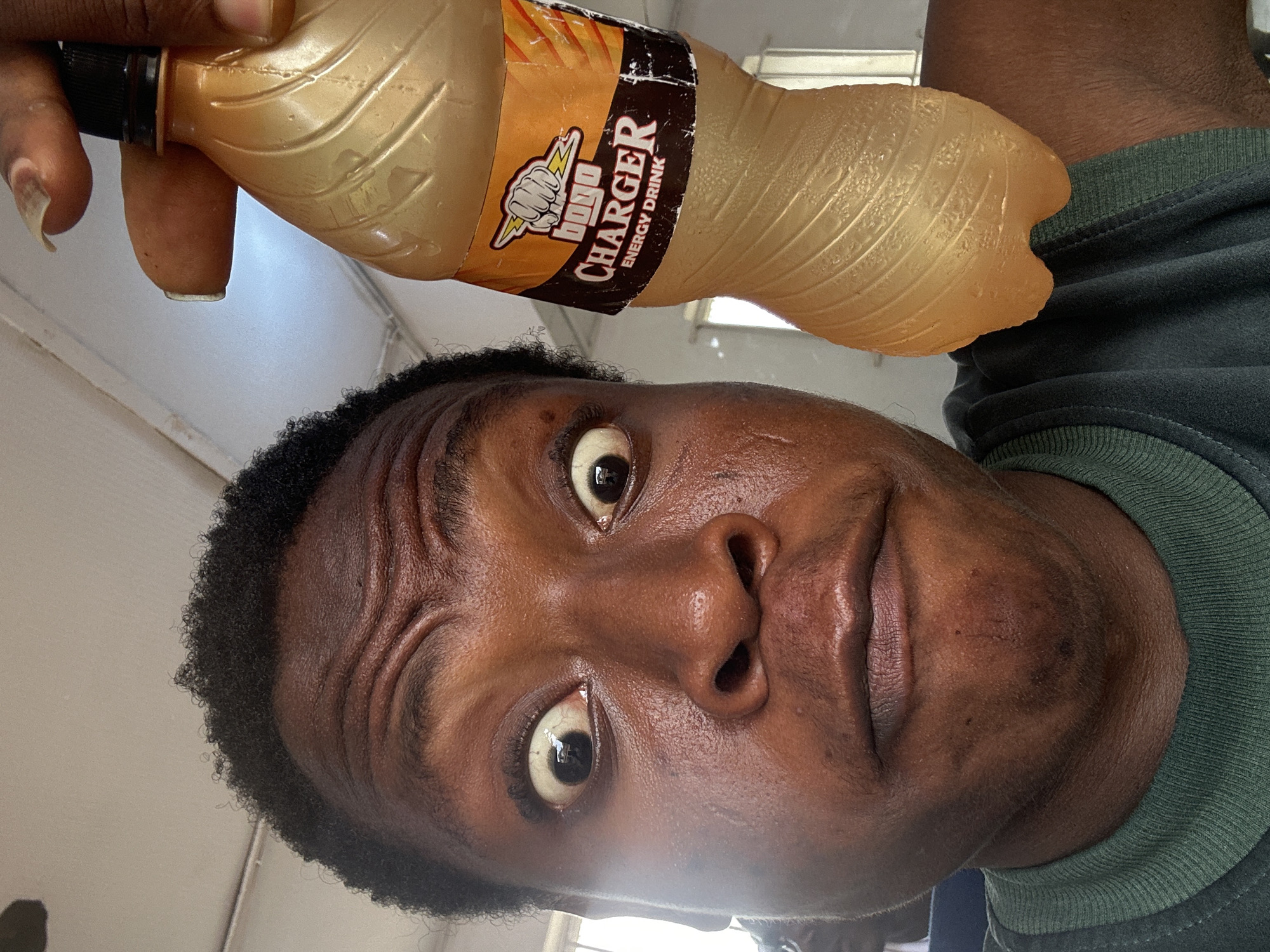 A dark-skinned male with short black hair, wrinkles on his forehead, and wide eyes holds a clear plastic bottle of "Charger" energy drink. The bottle is in his right hand. He wears a dark green shirt with a green neckband. The location is Unknown, Unknown.