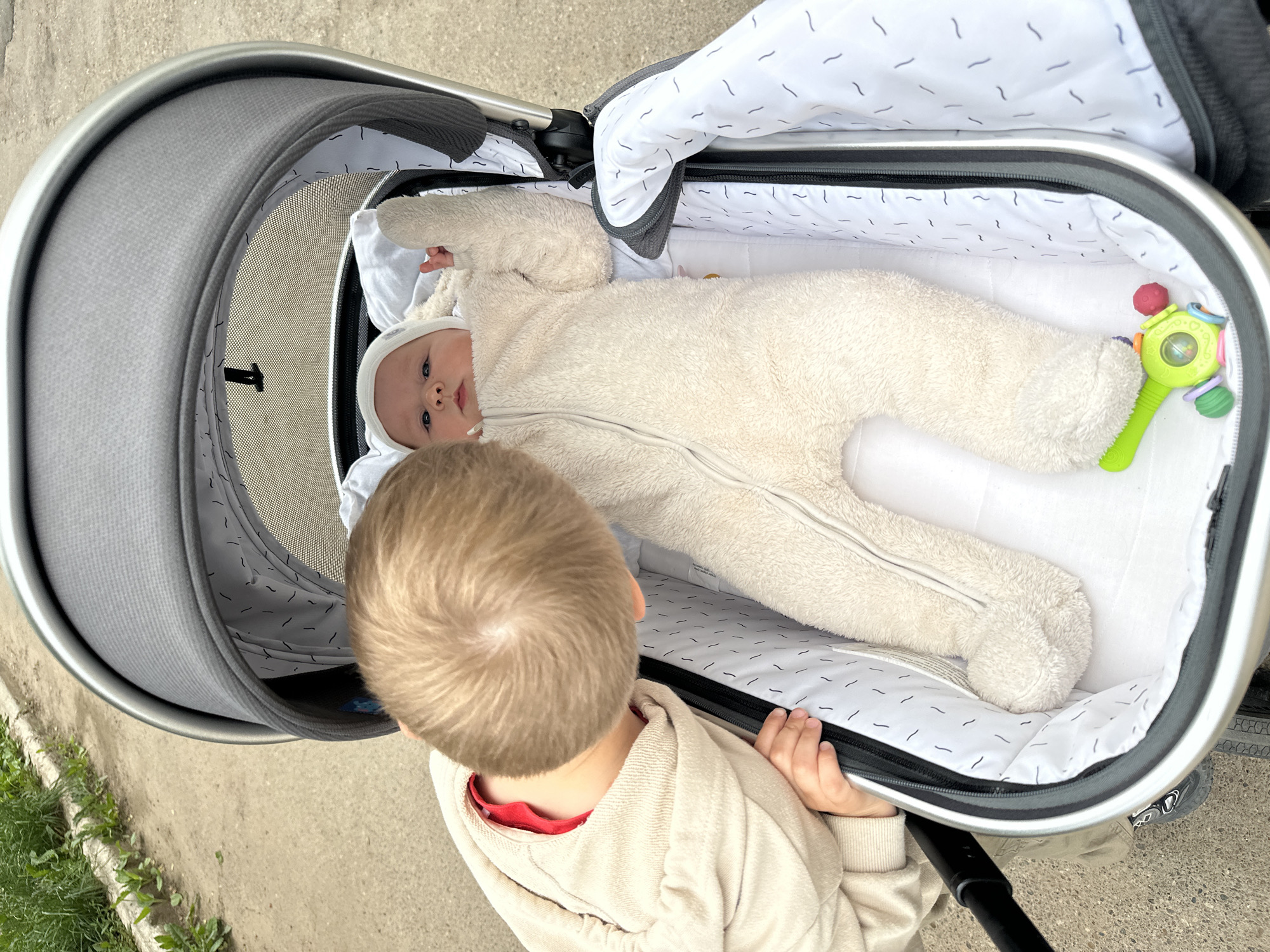 A blond-haired boy wearing a beige hoodie and a red undershirt leans over a baby in a stroller. The baby is wearing a cream-colored fleece snowsuit and a matching hat. A green and pink rattle lies in the stroller next to the baby. The stroller is parked on a concrete sidewalk next to a grassy area. The background shows a sidewalk and some vegetation. The scene appears to be outdoors in Chaikovsky, Russia, during daylight.